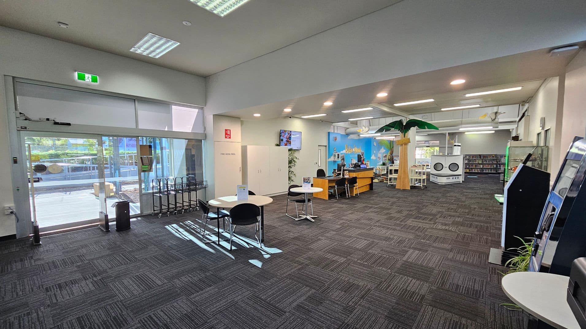 A spacious interior with a grey carpeted floor. On the left, there are large glass doors leading outside, with a row of trolleys nearby. In the centre, there are round tables with chairs. Towards the back, a reception desk is visible, with a colourful mural on the wall behind it. On the right, there are self-service machines and a display case. The ceiling is equipped with fluorescent lights, and a decorative palm tree stands near the centre. Bookshelves are visible in the background.