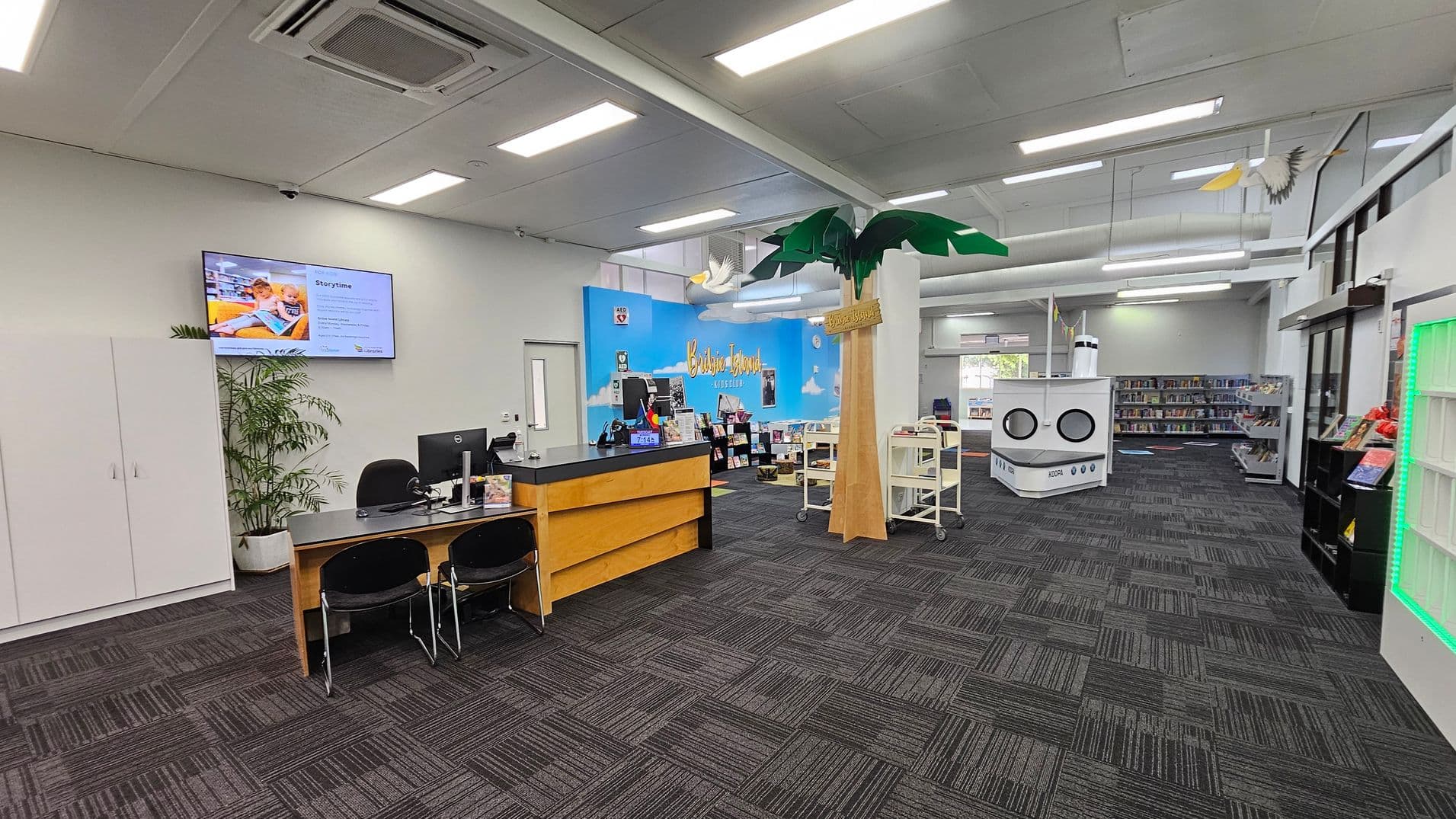 A library interior with a grey carpeted floor and a white ceiling with fluorescent lights. On the left, there's a reception desk with computers and chairs, and a potted plant nearby. A wall-mounted screen displays information. In the centre, a large decorative palm tree stands next to a blue wall with "Bribie Island" written on it. Bookshelves and a white cylindrical structure resembling a ship are visible in the background. On the right, more bookshelves and a display with green lighting.