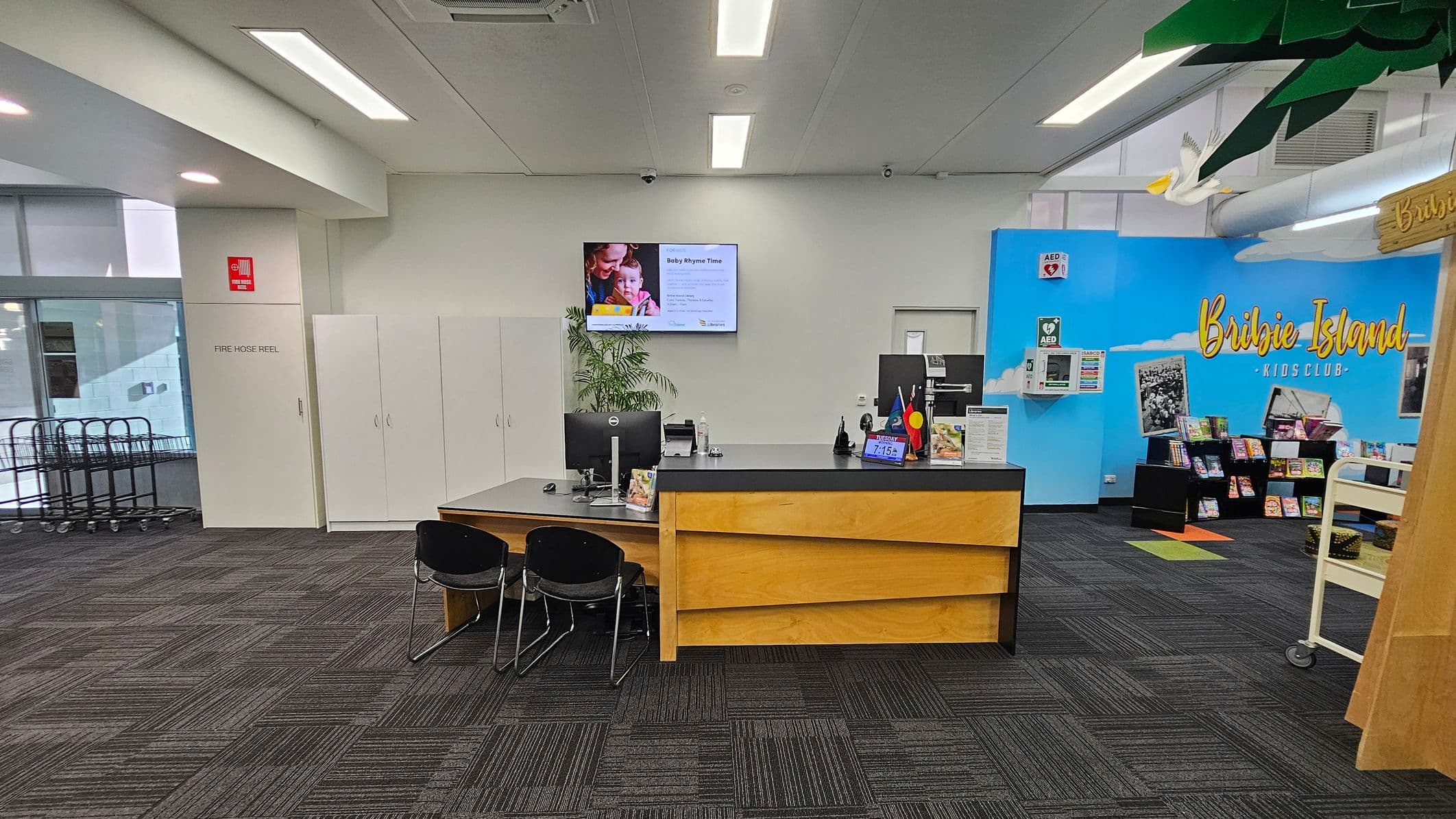 A library interior with a reception desk in the centre, featuring two black chairs and computer monitors. Behind the desk, a wall-mounted screen displays information. To the left, a fire hose reel is marked on a white door, with trolleys near a window. On the right, a blue wall with "Bribie Island Kids Club" text and a display of books. The ceiling has fluorescent lights, and the floor is covered with dark patterned carpet tiles. A white bird decoration hangs from the ceiling.