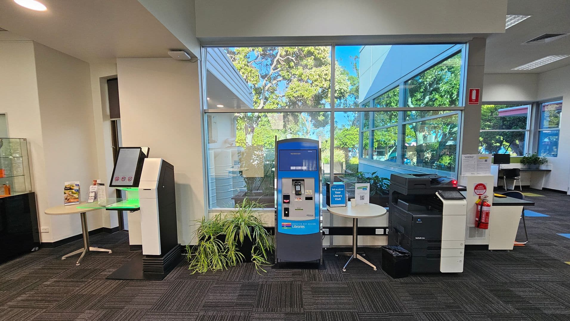 A room with a large window in the background showing trees outside. On the left, there's a kiosk with a screen and a small round table with brochures. In the centre, a blue and grey machine is flanked by potted plants. On the right, there's a printer with a fire extinguisher nearby. The floor is carpeted with a dark pattern, and the ceiling has recessed lighting.
