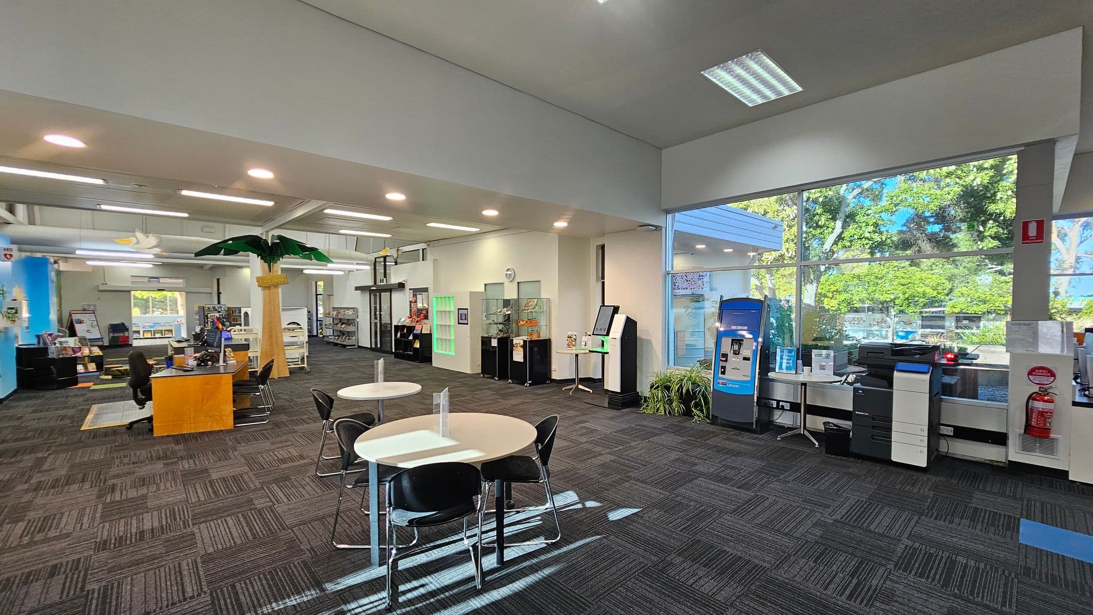 A spacious interior with a high ceiling and large windows on the right, allowing natural light to illuminate the room. In the foreground, there are round tables with black chairs. To the right, a blue kiosk and a printer are near a fire extinguisher. The left side features a reception desk with chairs and a decorative palm tree. Bookshelves and display cases line the walls, and the floor is covered with patterned carpet tiles.