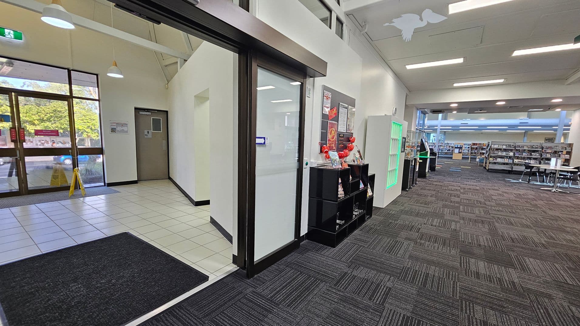 On the left, there is a glass entrance door with a tiled floor and a caution sign. A door is visible on the adjacent wall. In the centre, a glass door leads into a carpeted area with a black shelving unit displaying brochures and a bulletin board above. On the right, the space opens into a library with bookshelves, tables, and chairs. The ceiling has fluorescent lights, and a green-lit display case is near the entrance.
