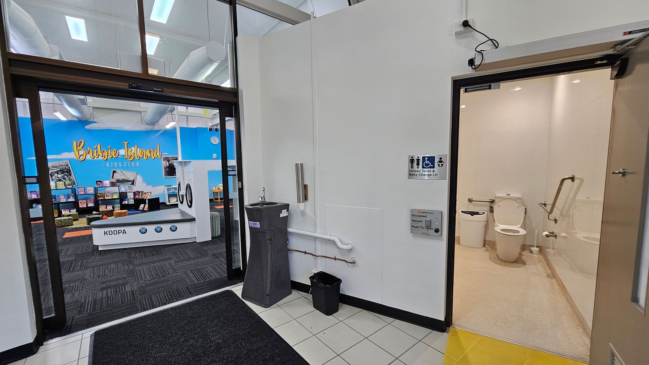 A hallway with a black mat on a tiled floor. On the left, there's an entrance to a room with a blue wall displaying "Bribie Island Kids Club" and shelves filled with books. On the right, a door leads to a unisex toilet with a baby change facility, featuring a white toilet and handrails. A grey water fountain and a small black bin are mounted on the wall outside the toilet. The ceiling has visible ducts and lights.