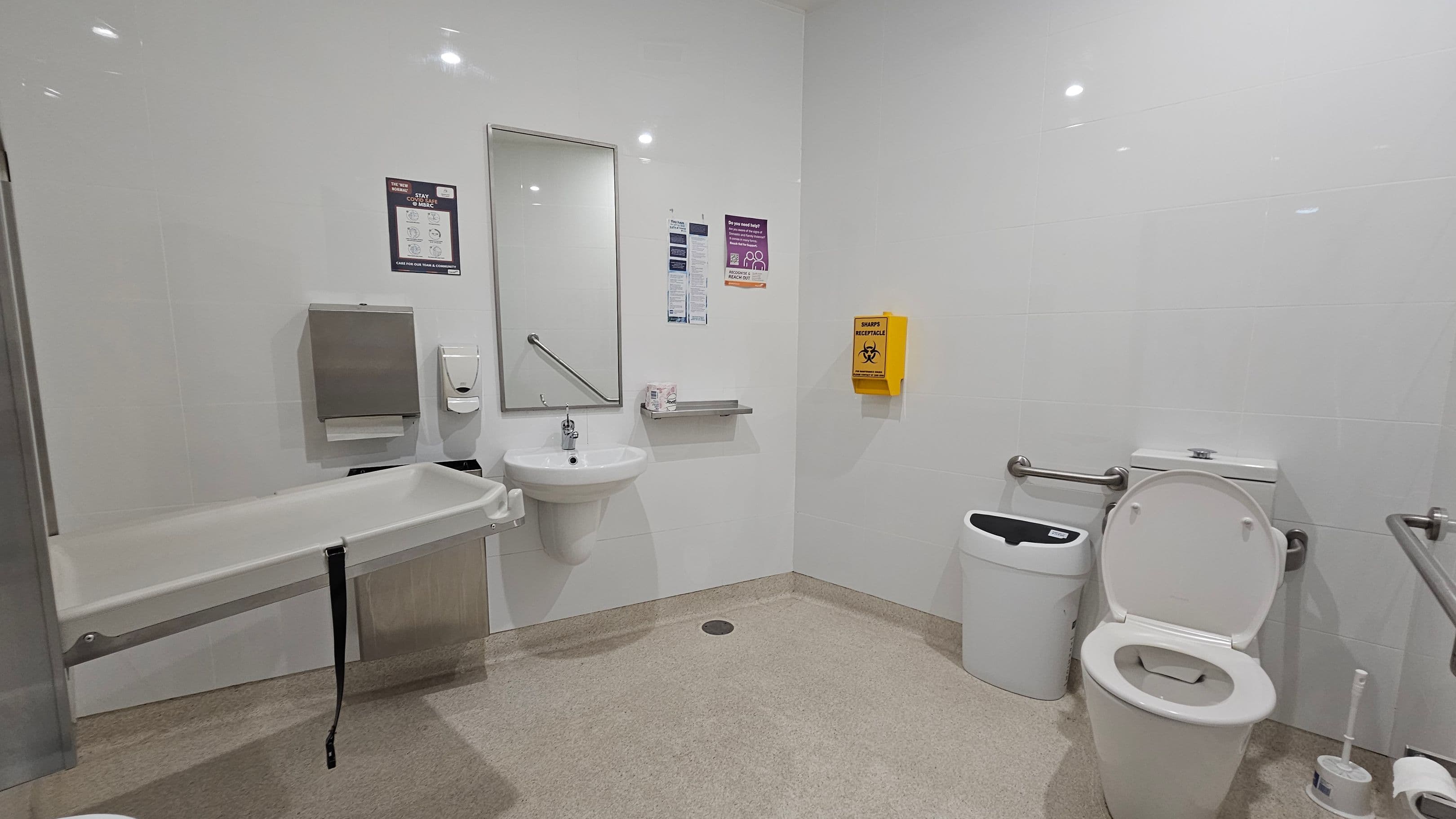 A clean, accessible bathroom with white walls and a light-coloured floor. On the left, a foldable changing table is mounted on the wall. In the centre, a sink with a mirror above it is flanked by a paper towel dispenser and a soap dispenser. A yellow sharps disposal box is mounted on the wall. On the right, a toilet with support rails is next to a bin and a toilet brush. The ceiling has recessed lighting.