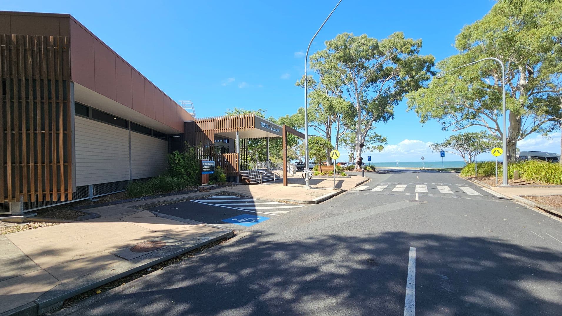 A street scene with a building on the left featuring vertical wooden slats and a sign reading "Seafood Market". A pedestrian crossing leads towards the sea in the background. Trees line the right side of the street, providing shade. A blue disabled parking space is visible in the foreground. The sky is clear and blue, and there are street lamps along the road.