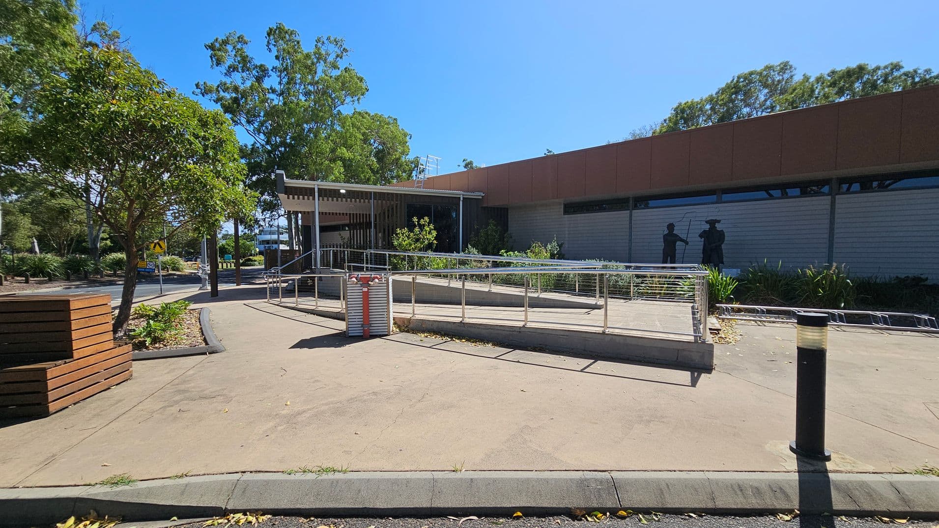 A paved area with a metal railing ramp in the centre. To the right, a building with a red-brown facade and a mural of two figures. On the left, a tree and a wooden bench. The background features more trees and a road with a pedestrian crossing sign. The ground is concrete, and there are a few scattered leaves. A black bollard is visible in the foreground.