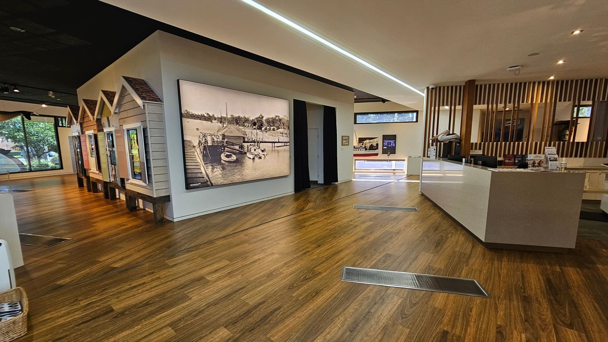 Interior of a modern exhibition space with wooden flooring. On the left, there are small house-like structures with displays and a large historical photograph of a dock scene. The right side features a sleek, white reception desk with informational materials. Behind the desk, vertical wooden slats create a partition. Large windows in the background allow natural light to enter, and the ceiling has recessed lighting.