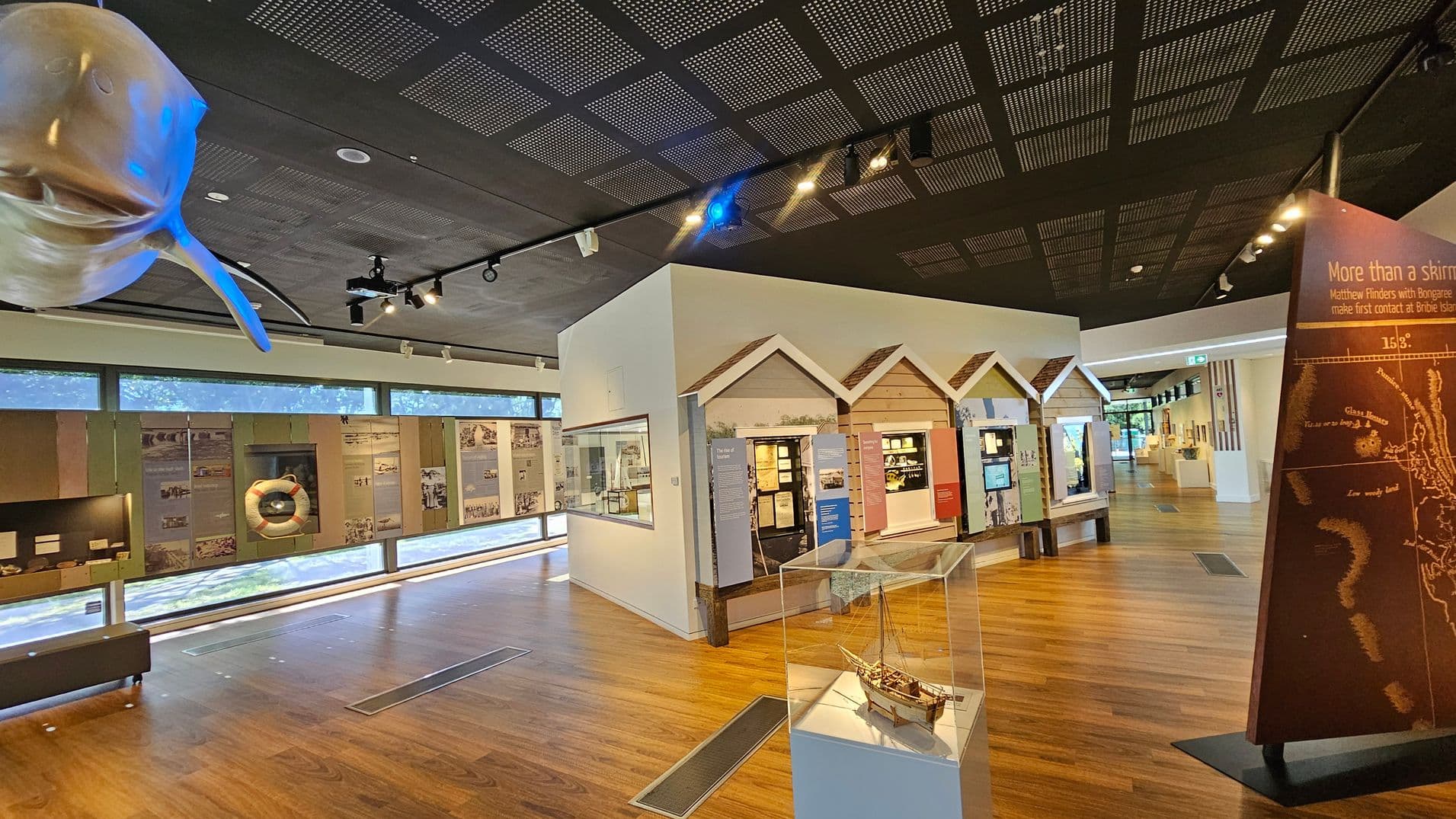A museum interior with wooden flooring and a dark, perforated ceiling. On the left, a large model of a fish is suspended from the ceiling. The background features a long wall with large windows and various historical displays, including a life preserver. In the centre, a glass case contains a model ship. On the right, an angled display board with text and a map. The space is well-lit with track lighting, and there are informational panels and exhibits throughout.