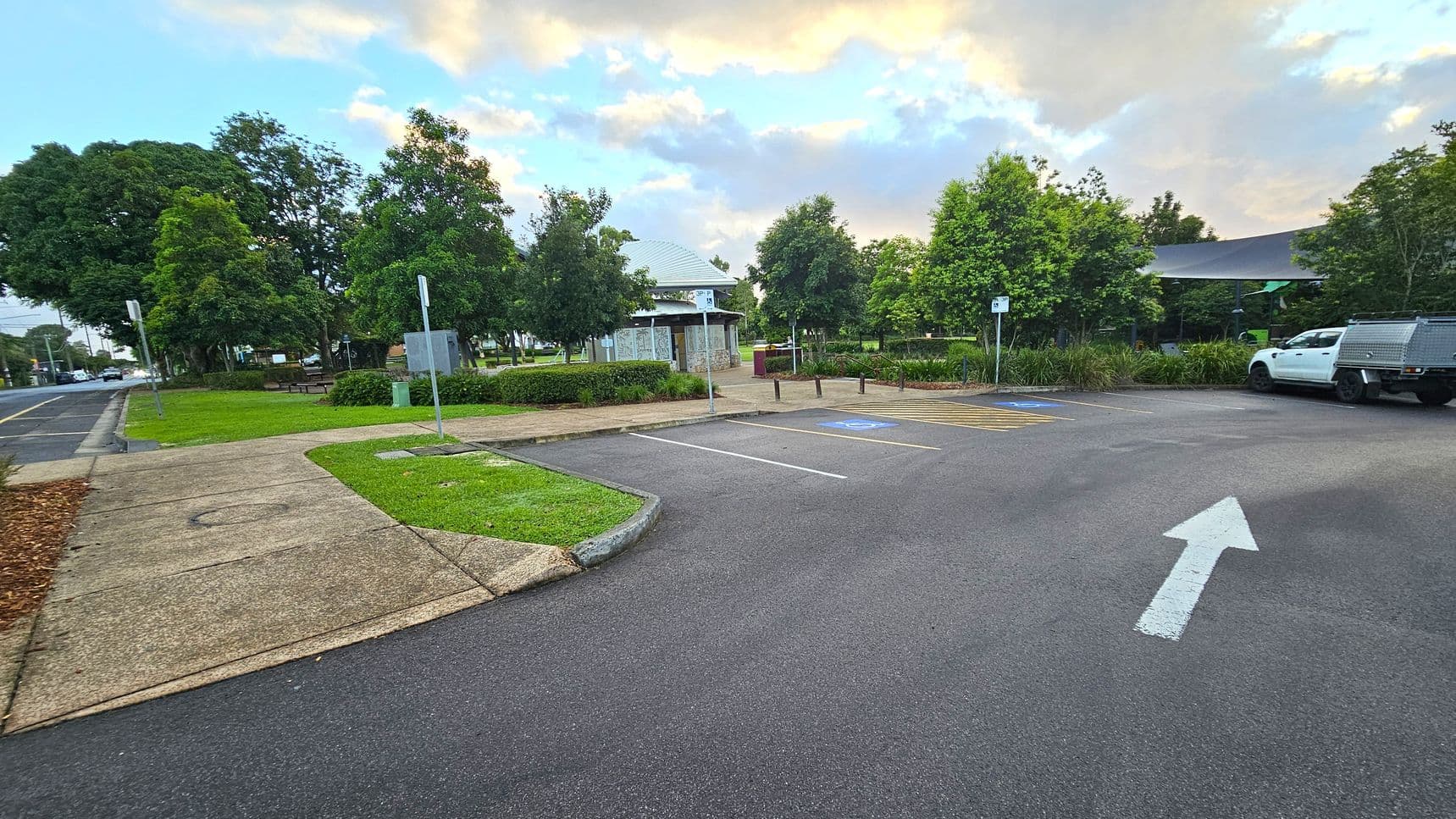 A paved parking area with marked spaces, including a disabled parking spot, is in the foreground. An arrow is painted on the asphalt, indicating traffic direction. To the right, a white utility vehicle is parked. In the background, there are several trees and a small building with a metal roof. A footpath runs along the left side, bordered by grass and leading to a street. The area is surrounded by greenery, including shrubs and more trees.