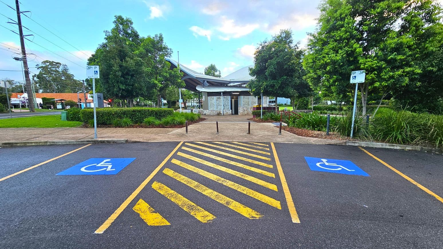 In the foreground, there are two parking spaces marked with blue wheelchair symbols, separated by a yellow-striped area. Signs indicating "3P" parking are visible above each space. The background features a pathway leading to a building with a modern, curved roof. Surrounding the path are green shrubs and trees, with a grassy area to the left. The pavement is dark, and the sky is partly cloudy.