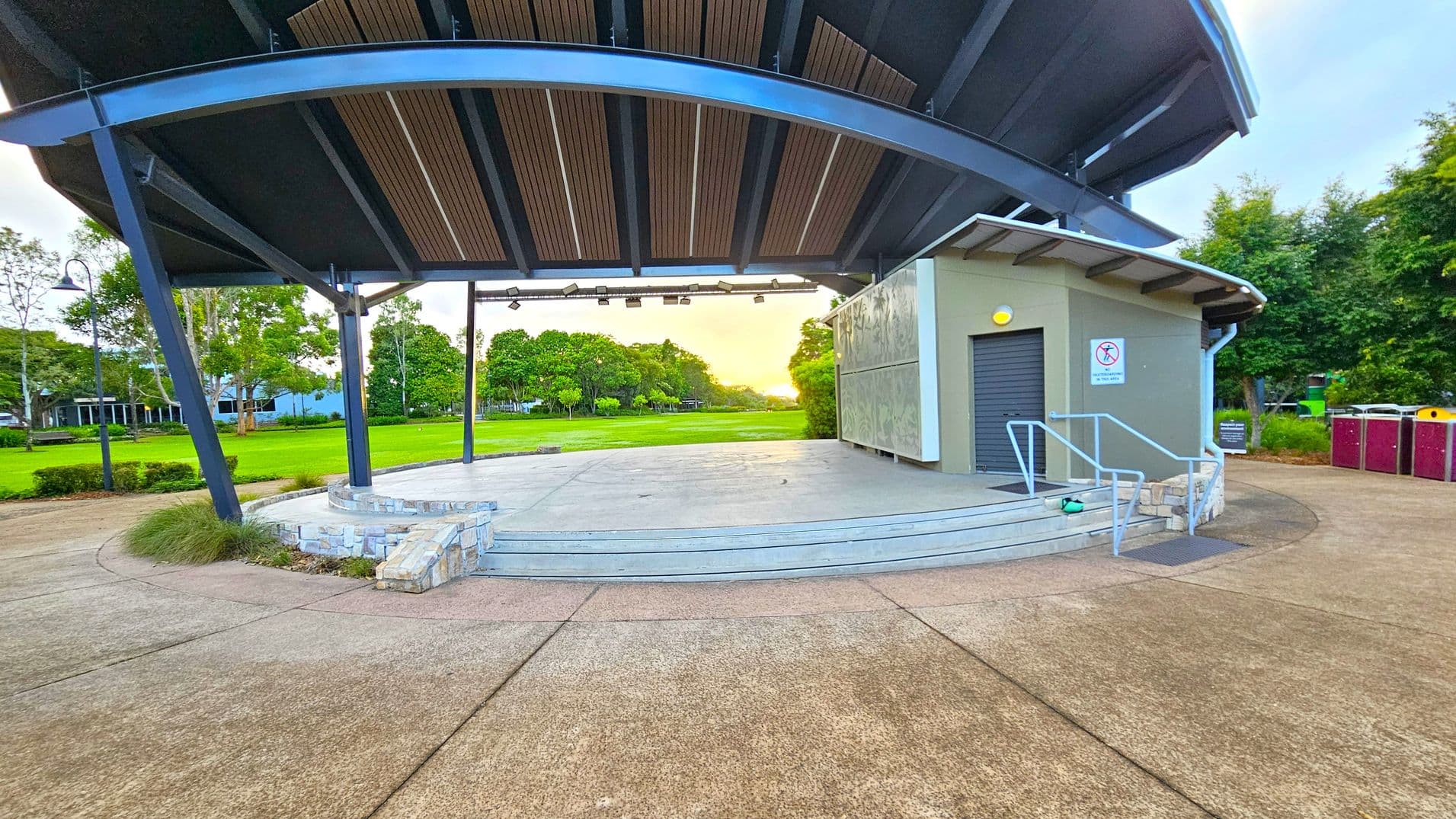 A covered outdoor stage with a curved metal roof and wooden slats. The stage has a stone base and steps leading up to it. On the right, there's a small building with a closed roller door and a sign prohibiting smoking. Metal railings are present beside the steps. In the background, there's a grassy field bordered by trees. The foreground features a concrete pavement with a few patches of grass.