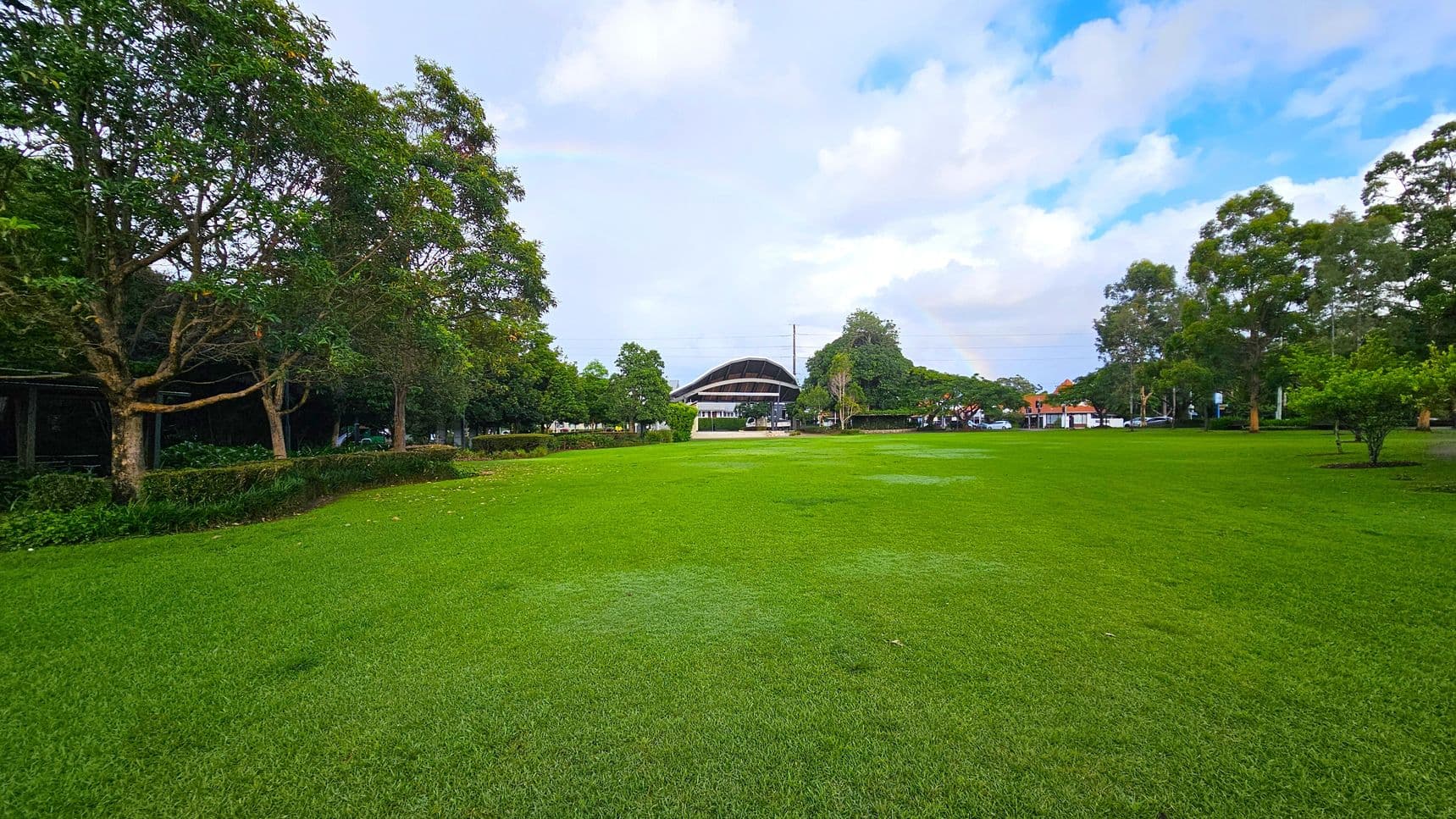 A large, open grassy field stretches across the foreground. On the left, there are several trees with dense foliage, and a hedge runs parallel to them. In the centre background, a modern, curved-roof structure is visible, surrounded by more trees. To the right, additional trees and a few parked cars can be seen. The scene is well-lit, suggesting daytime.