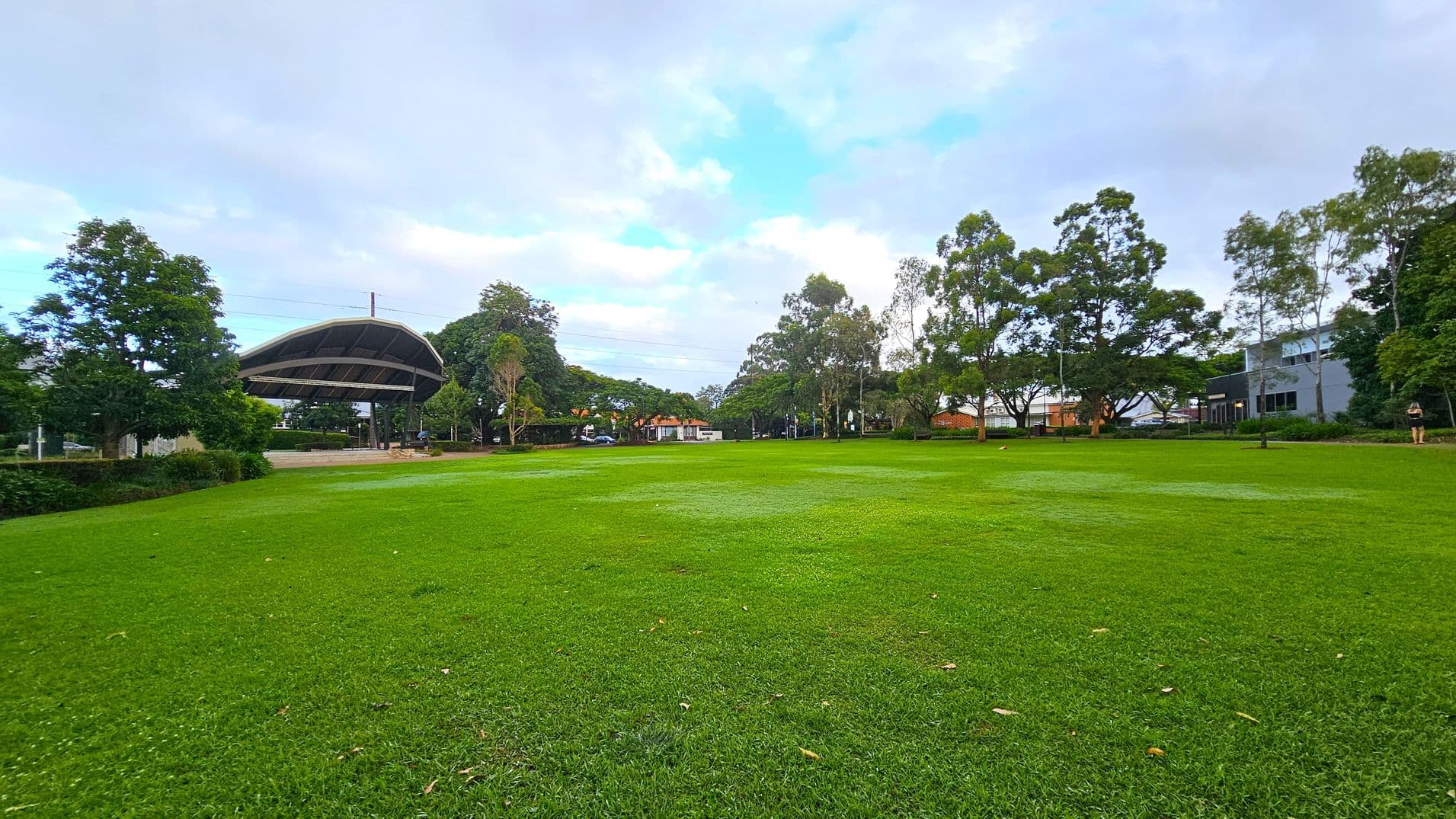 A large grassy field stretches across the foreground, with a few scattered leaves. In the background, there are several tall trees and a covered structure to the left, resembling a pavilion. Buildings are visible among the trees on the right side. The area is open and spacious, with a few people walking in the distance.