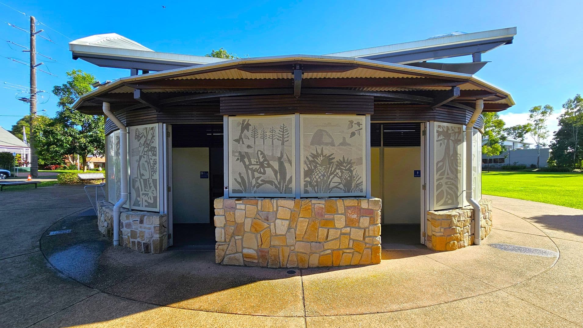 Circular public restroom with a stone base and decorative panels featuring nature motifs. The roof is corrugated metal with a central peak. The entrance is open, revealing a dark interior. Surrounding the structure is a paved area, with grass and trees in the background. A power pole and a building are visible to the left. The sky is clear and blue.