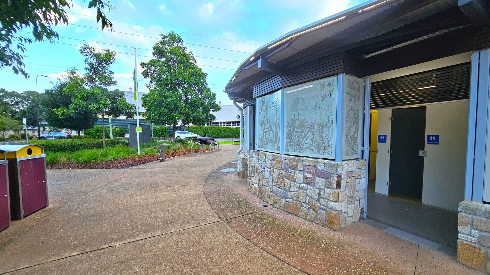 A public restroom facility is on the right, featuring stone and metal construction with open doors displaying male and female signs. The path curves around the building, leading to a landscaped area with trees and shrubs on the left. A bench and a flagpole are visible in the background, along with parked cars and a building. The ground is paved with concrete, and there are waste bins on the left.