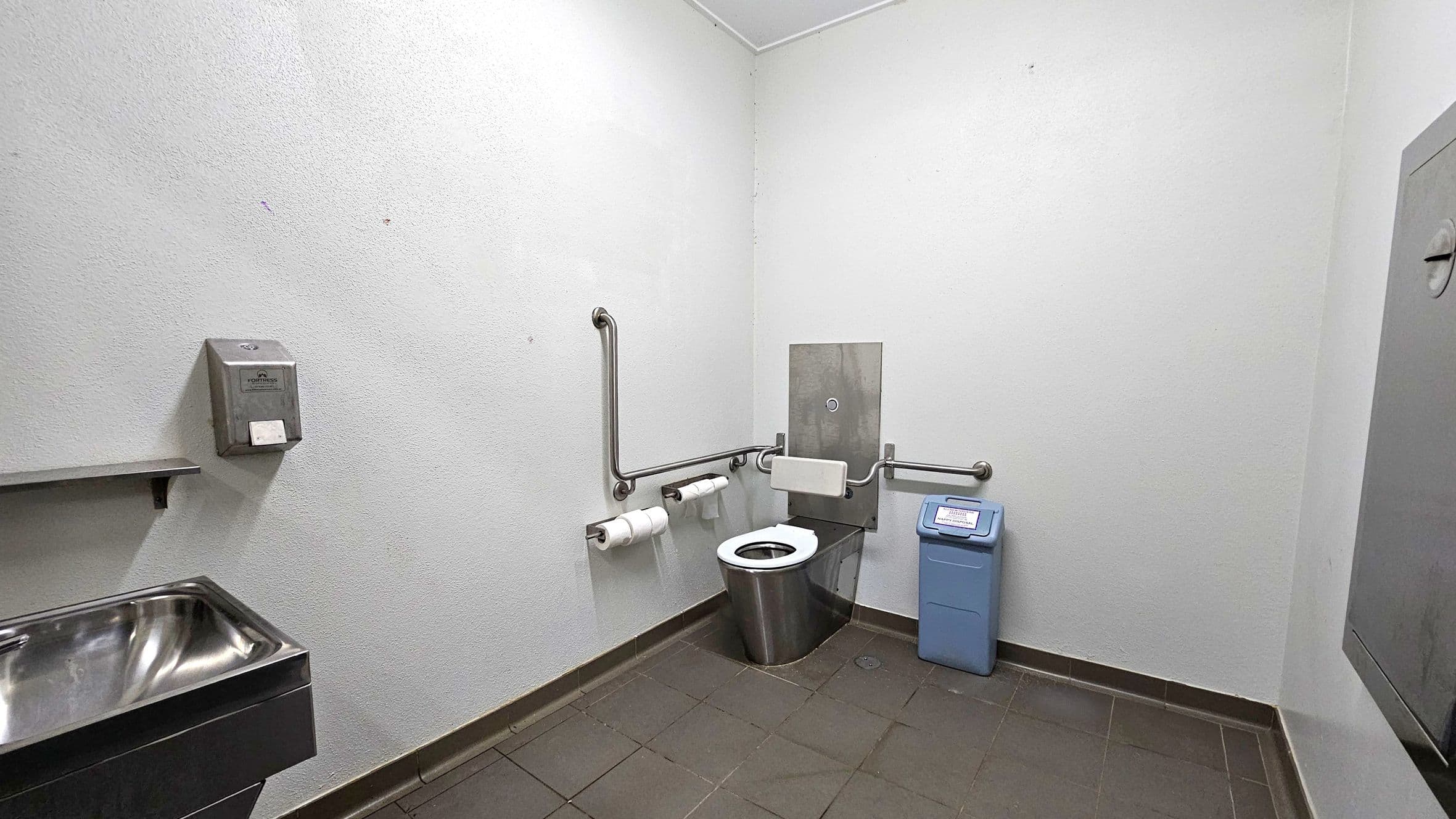 A public restroom with a stainless steel toilet in the centre, equipped with grab bars on both sides. To the right, a blue sanitary bin is placed on the tiled floor. On the left, a stainless steel sink is mounted on the wall, with a soap dispenser above it. The walls are plain and light-coloured, and the floor is covered with dark tiles. The ceiling is plain and light-coloured.