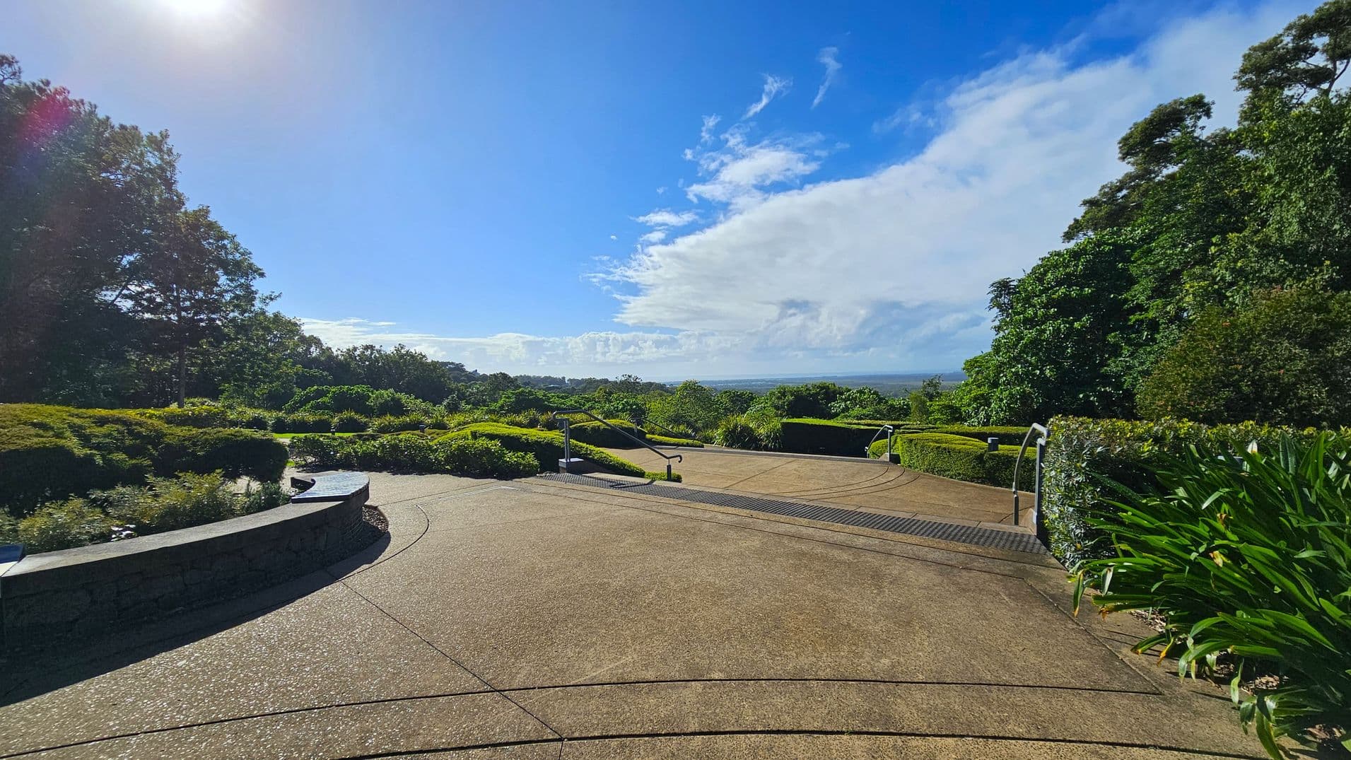 A paved pathway leads into a lush garden area. On the left, there are dense trees and a curved stone bench surrounded by shrubs. On the right, more greenery and manicured bushes line the path. In the centre, the path descends with metal railings on either side. The background features a distant view of the horizon under a partly cloudy sky. The sun is visible in the top left corner, casting light across the scene.