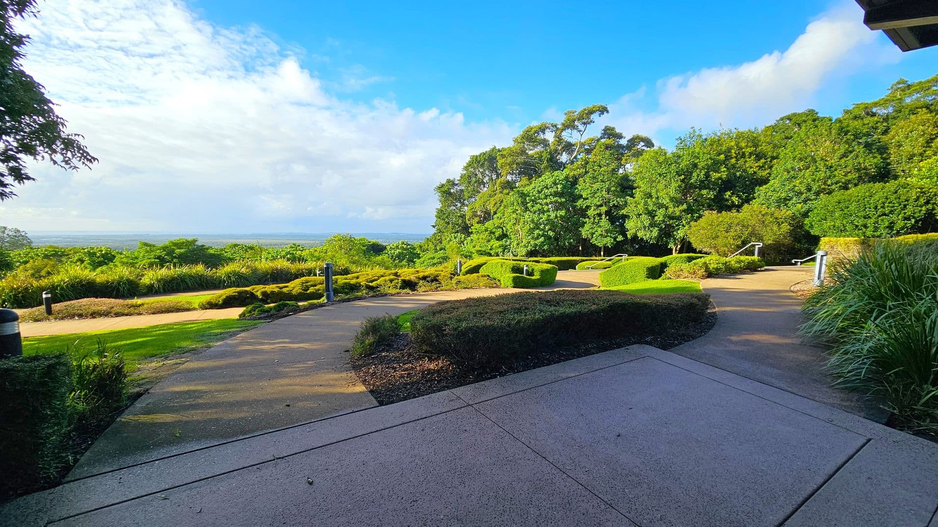 A paved pathway curves through a landscaped garden with neatly trimmed hedges and lush green trees on the right. On the left, there are more shrubs and a view of the distant horizon. The foreground features a textured concrete surface, while the background is filled with dense foliage and a clear view of the sky. Small bollard lights line the path, and there are handrails along the steps leading up the path.
