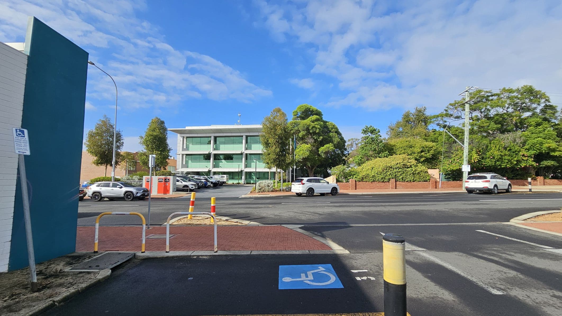 A street scene with a three-storey building in the centre, featuring large glass windows. In front of the building, there are several parked cars and trees. On the left, a blue wall with a disabled parking sign is visible. The foreground shows a disabled parking space marked on the ground. On the right, a white car is driving past, with more trees and a brick wall in the background. The road is bordered by a red brick pavement.