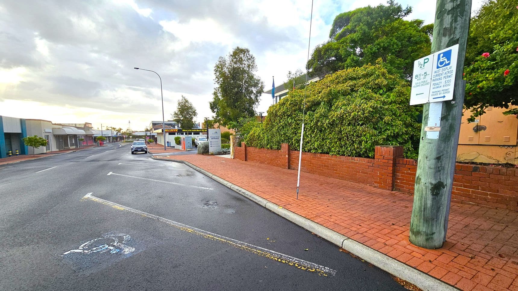 A street scene with a wet road and a single parked car on the left. The pavement is red brick, bordered by a low red brick wall with dense green foliage behind it. A wooden utility pole on the right has a sign indicating disabled parking. Buildings with shopfronts line the left side of the street, and a tall streetlamp is visible. The sky is overcast, and the area appears quiet.