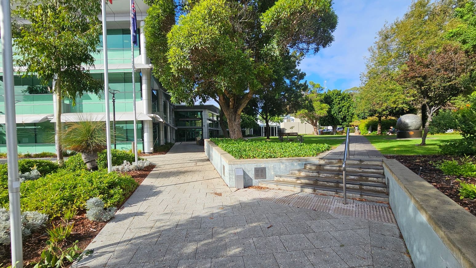 A modern building with large glass windows is on the left, partially obscured by trees and flagpoles. A paved walkway leads towards the entrance, flanked by landscaped greenery. In the centre, a large tree provides shade over a raised garden bed. On the right, a set of steps with a metal railing ascends to a grassy area with more trees and a large spherical sculpture. The ground is paved with grey tiles, and the area is well-maintained with various plants.