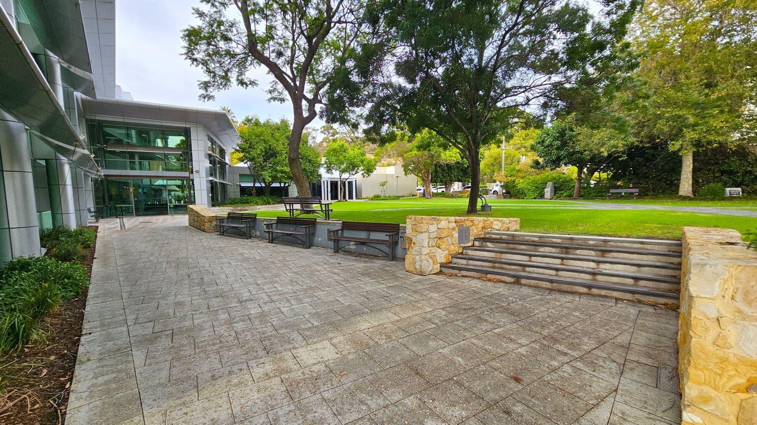 A modern building with large glass windows is on the left, adjacent to a paved walkway. In the centre, there are stone steps leading up to a grassy area with trees and several benches. The right side features more greenery and a stone wall. The background includes additional trees and a small structure, with a few parked vehicles visible.