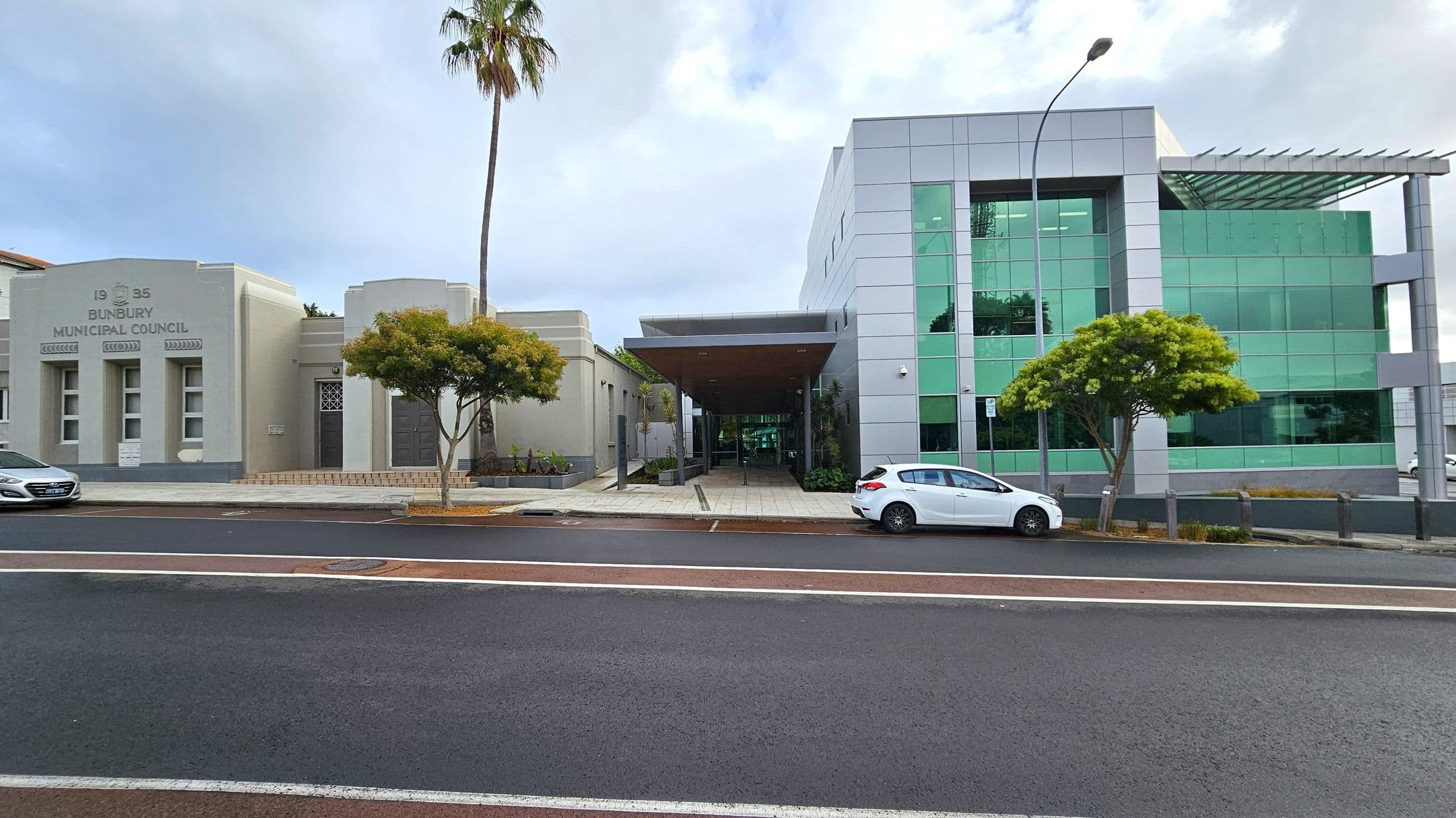 On the left, a beige building with "1935 Bunbury Municipal Council" inscribed above three windows. A palm tree and a smaller tree are in front. In the centre, a covered walkway leads to a modern building on the right, featuring green-tinted glass panels. A white car is parked in front of this building, and a small tree is nearby. The foreground shows a paved road with white and red markings.