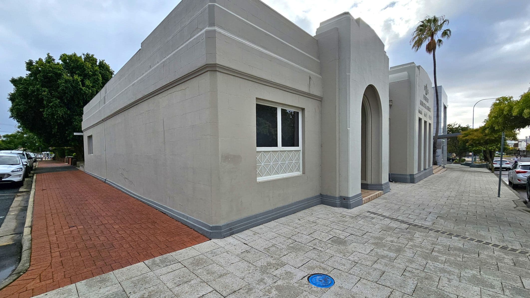 A grey building with an arched entrance and rectangular windows is on the right. The pavement is made of light grey bricks, with a blue circular cover visible. A palm tree and other trees line the street, with parked cars along the kerb. On the left, a red brick path runs alongside the building, leading to more parked cars and greenery.