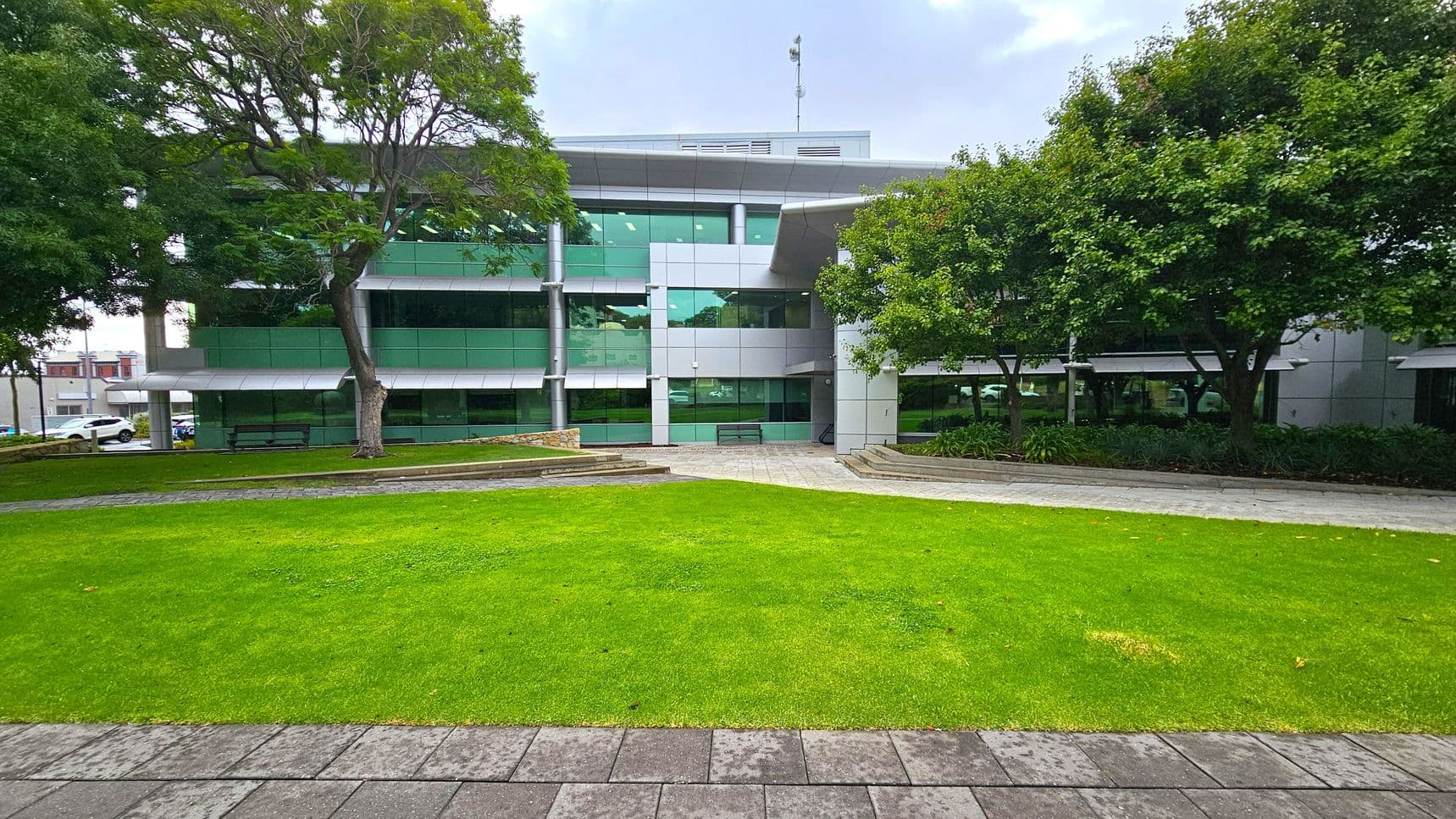 A modern building with large green-tinted windows and a grey facade is in the background. In the foreground, there is a well-maintained lawn bordered by a paved walkway. Trees with dense foliage are on both sides, providing shade. Benches are placed near the building, and there are landscaped areas with plants. The scene is calm and organised, with a mix of natural and architectural elements.