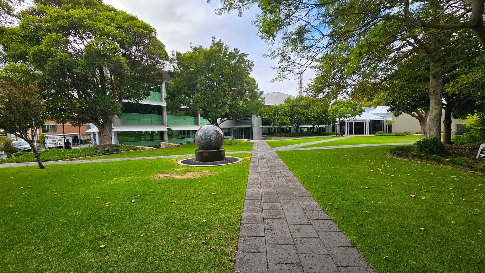 A paved path leads through a grassy area towards a large spherical sculpture on a pedestal in the centre. Surrounding the sculpture are well-maintained lawns with scattered fallen leaves. On the left, a large tree provides shade near a modern building with green-tinted windows. On the right, more trees and another building with a white facade are visible. The background features additional trees and structures, creating a serene park-like setting.