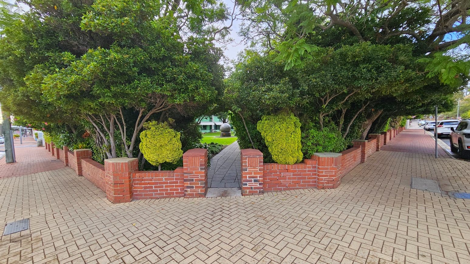 A brick pathway leads through a small entrance flanked by red brick walls and dense green foliage. The path continues under an arch formed by trees and shrubs, leading to a grassy area with a large spherical sculpture in the distance. The foreground features a herringbone-patterned brick pavement, with parked cars and a street visible on the right. The left side shows more brick walls and trees lining the pavement.