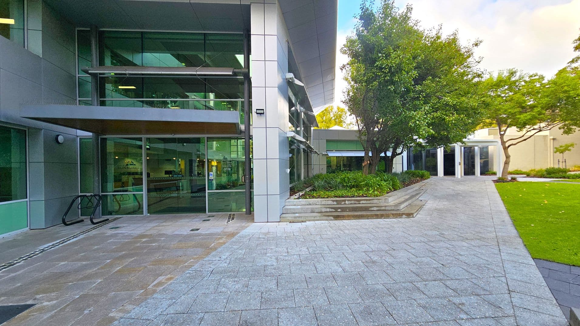 On the left, a modern building with large glass windows and a metal awning. A bike rack is visible near the entrance. In the centre, a paved walkway leads towards the building. On the right, a landscaped area with green grass, trees, and shrubs. A path extends towards another building entrance in the background. The scene is well-maintained and orderly.