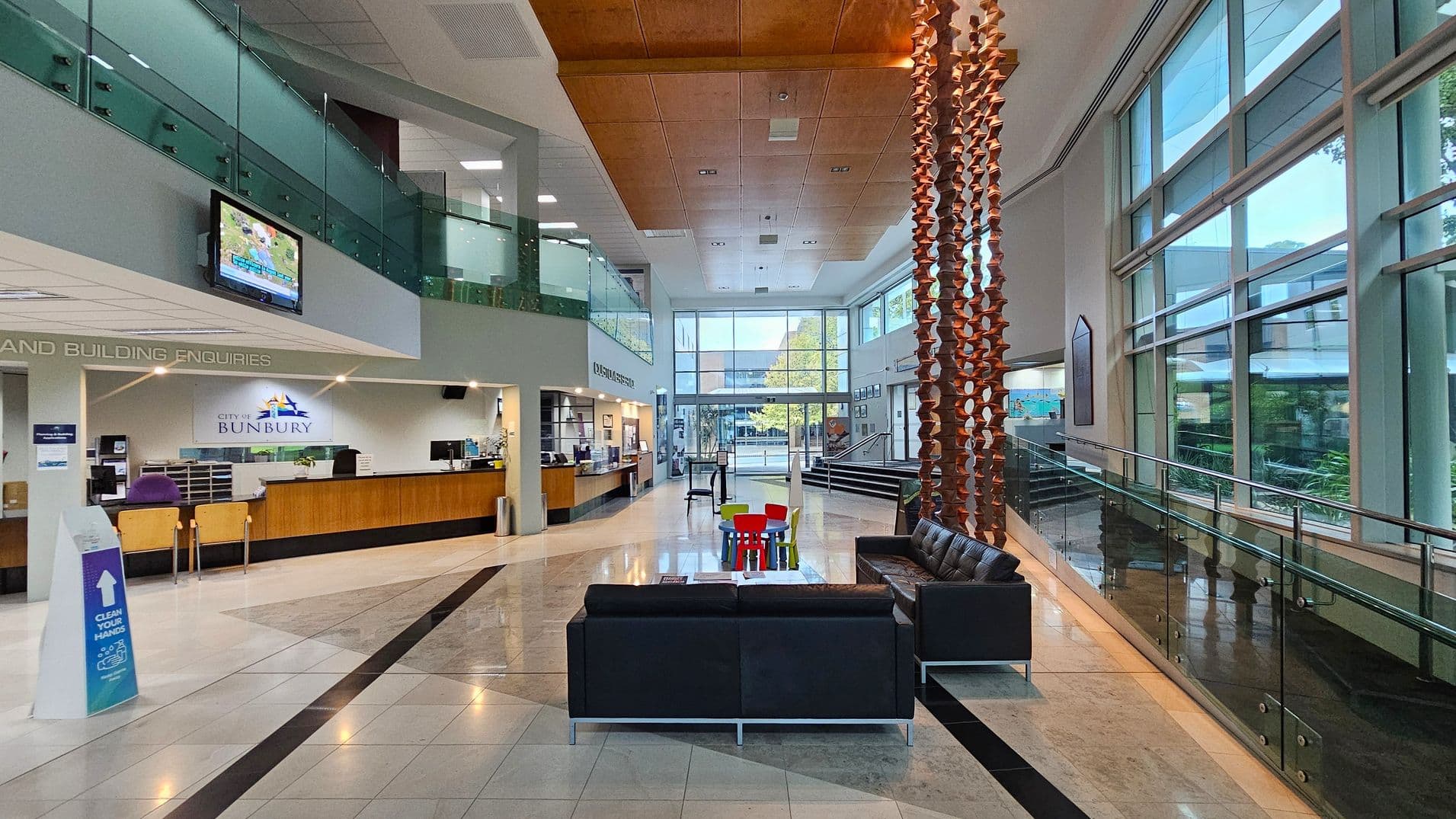 A spacious, modern lobby with a high ceiling and large windows on the right, allowing natural light to flood in. On the left, there's a reception desk with a sign reading "City of Bunbury" and a television mounted above. In the centre, a seating area with black sofas and a small table with colourful chairs. A decorative wooden sculpture hangs from the ceiling. The floor is polished with a geometric pattern, and a glass railing lines the right side.