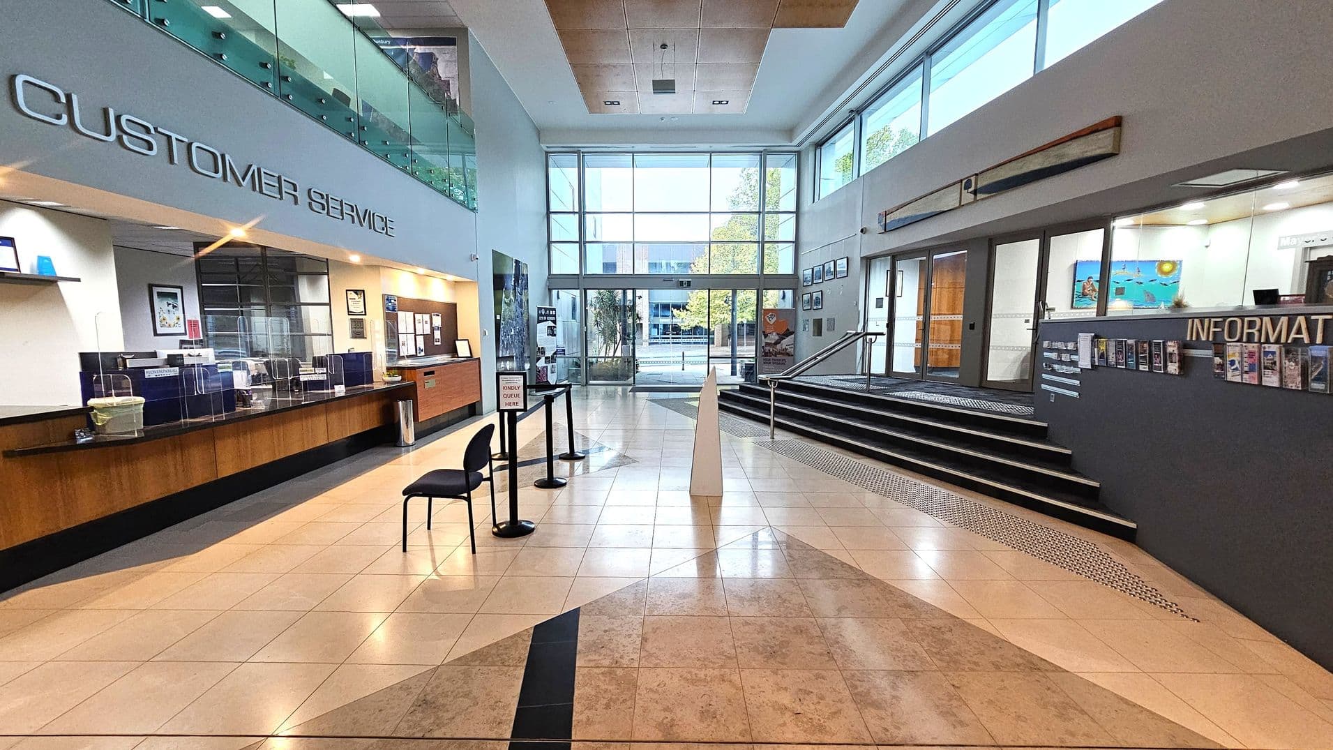 A spacious lobby with a tiled floor. On the left, a customer service area with a long wooden counter and protective screens. Above, a sign reads "CUSTOMER SERVICE". Two chairs are placed near a queue stand. On the right, an information desk with brochures and a sign. A staircase leads up to a glass entrance with large windows, allowing natural light to fill the space. The ceiling is high with recessed lighting.
