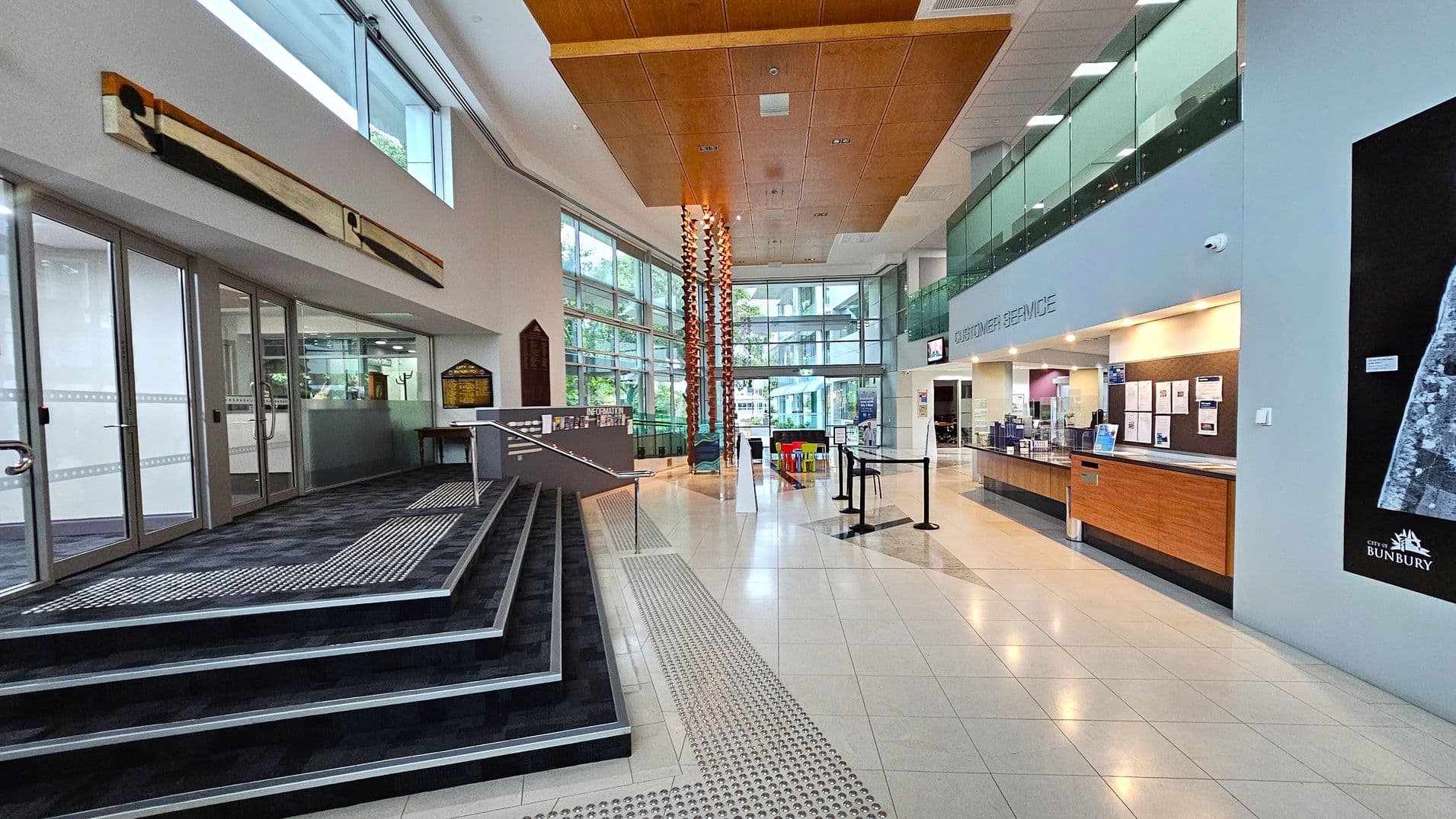 A spacious interior with a modern design. On the left, there are glass doors and a ramp with steps leading up. A long, abstract artwork is mounted on the wall above. In the centre, a tall, decorative column reaches up to the wooden ceiling. On the right, a customer service desk with a wooden counter is visible, along with a large map on the wall. The floor is tiled, and large windows in the background allow natural light to fill the space.