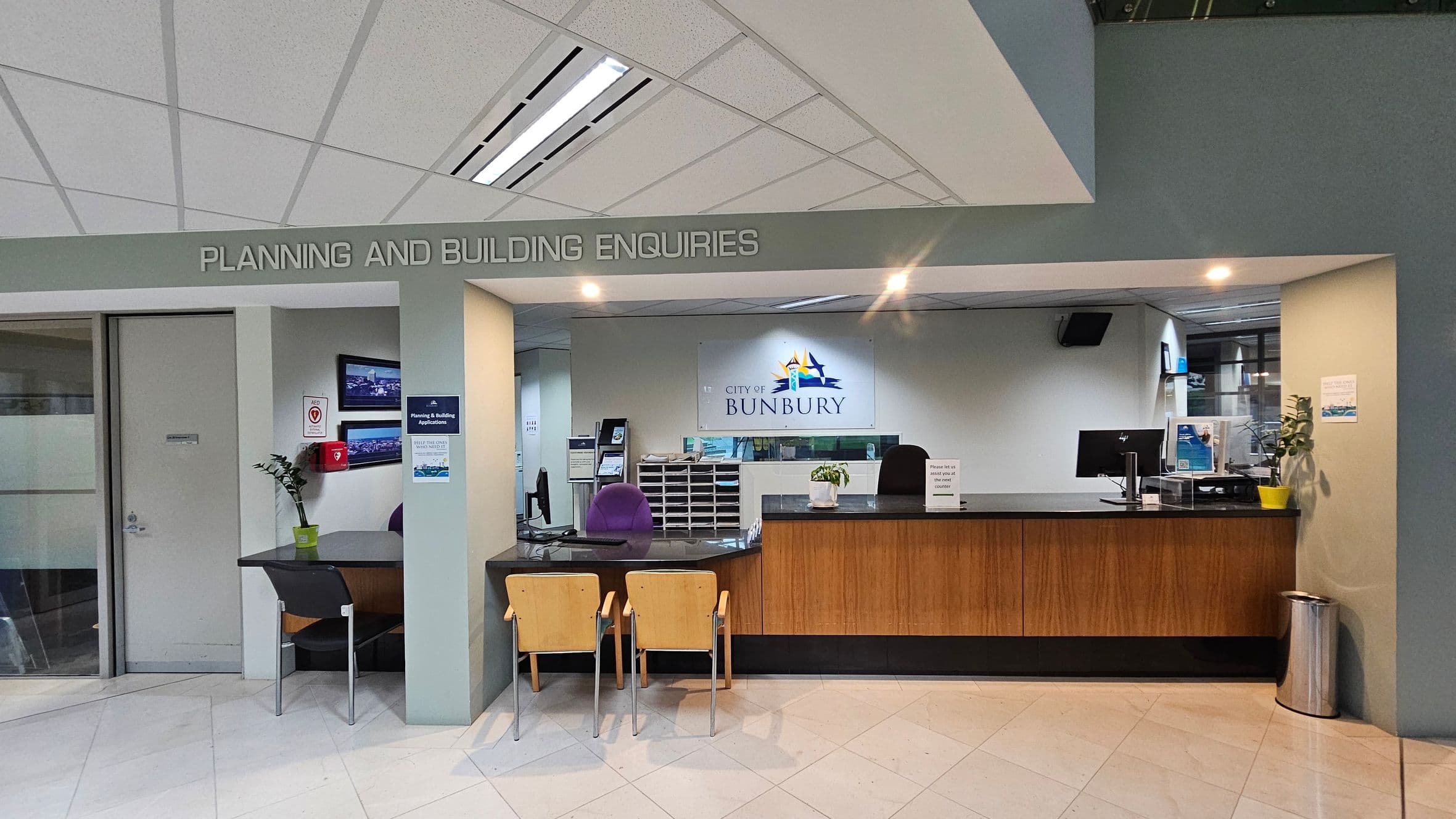 A reception area with a sign above reading "PLANNING AND BUILDING ENQUIRIES". On the left, there's a small table with a chair, a plant, and a wall-mounted screen. A door is adjacent to this setup. In the centre, a counter with two wooden chairs faces a desk with a computer and office supplies. Behind the counter, a sign reads "CITY OF BUNBURY". The floor is tiled, and the ceiling has a grid pattern with fluorescent lights. A small plant and a bin are on the right side of the counter.