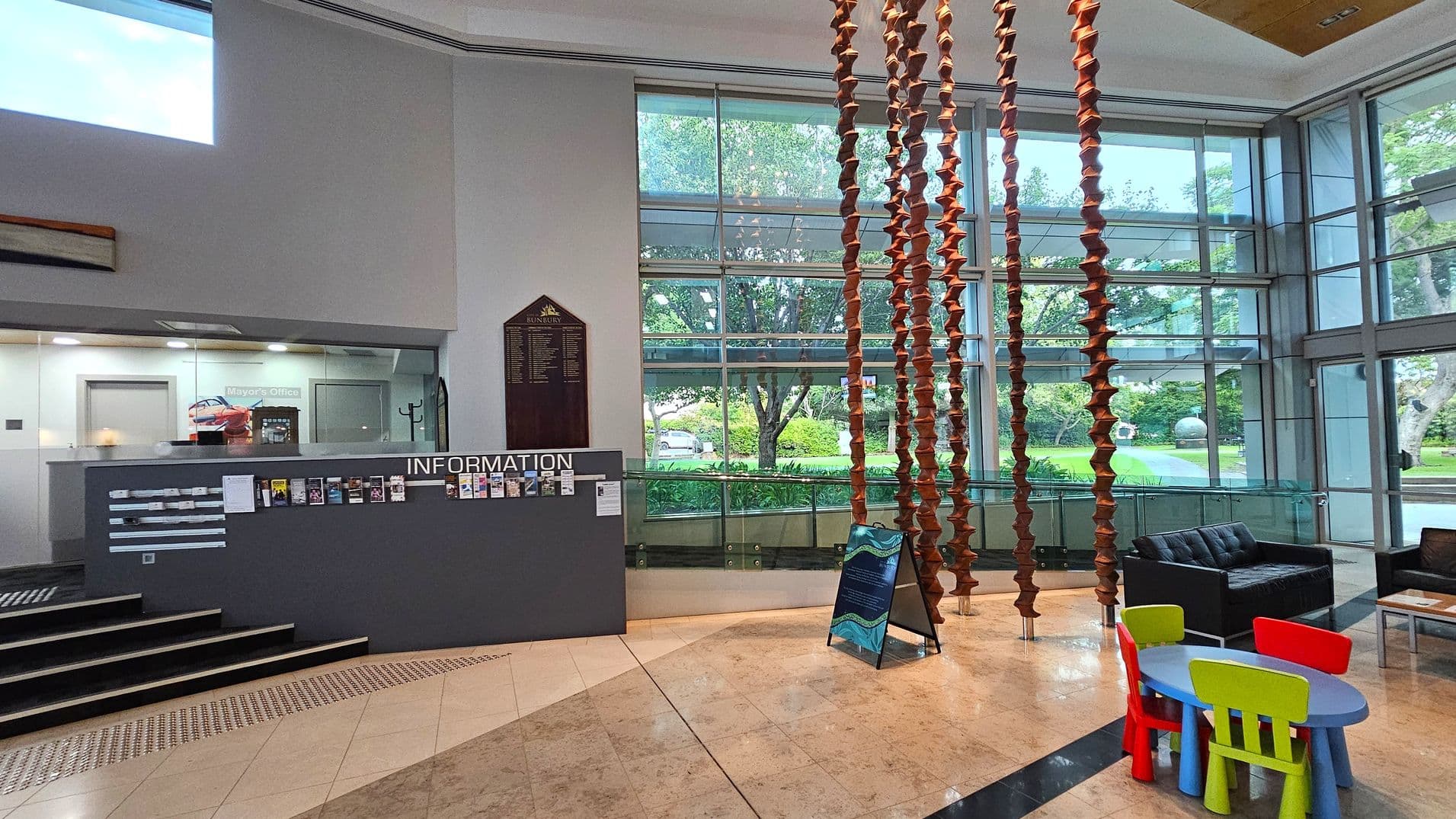 A spacious interior with a tiled floor and high ceiling. On the left, there's an information desk with brochures and a sign for the Mayor's Office. Behind the desk, a glass wall and door are visible. In the centre, a decorative hanging installation made of stacked, brown elements. On the right, a seating area with a black sofa and a small children's table with colourful chairs. Large windows in the background offer a view of greenery outside.