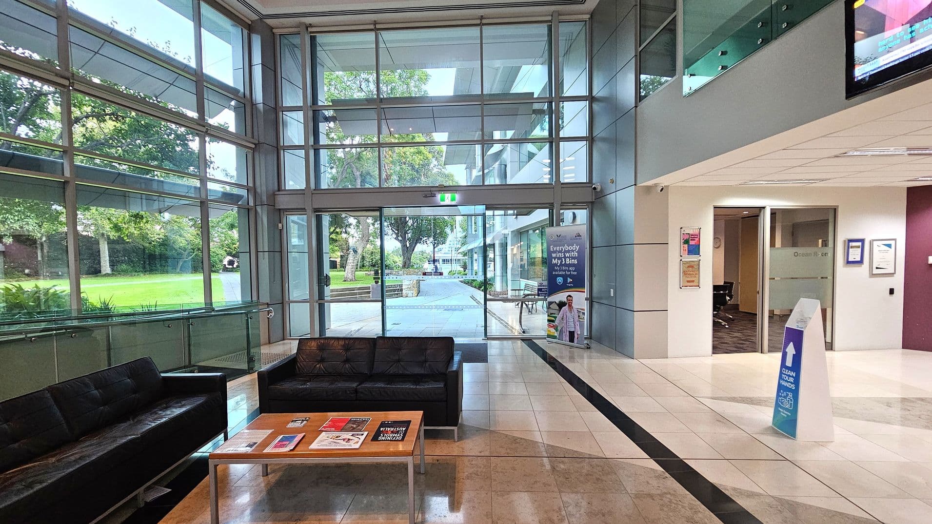 A modern lobby area with large glass windows on the left, offering a view of a green lawn and trees outside. In the centre, there is a glass entrance door with a safety exit sign above. To the right, a white wall with an open doorway leading to an office space. A black leather sofa and a wooden coffee table with magazines are in the foreground. The floor is tiled in a light colour with a dark strip, and a sign on the floor advises to clean hands.