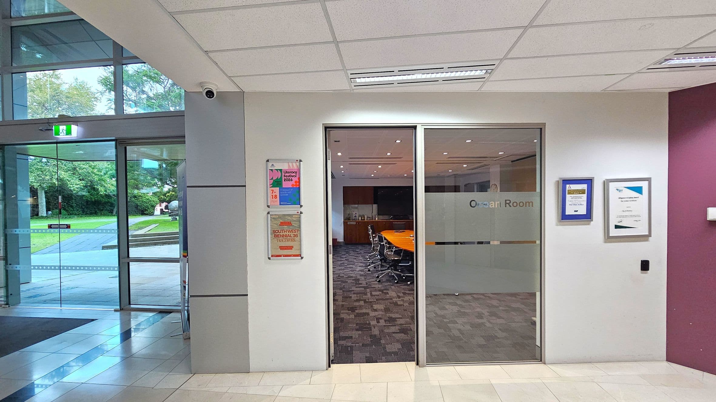 A hallway with a glass door on the left leading outside, showing a grassy area and trees. To the right, there is a glass entrance to a room labelled "Ocean Room," with a conference table and chairs visible inside. The wall beside the door has posters and certificates displayed. The floor is tiled, and the ceiling has recessed lighting and a security camera.