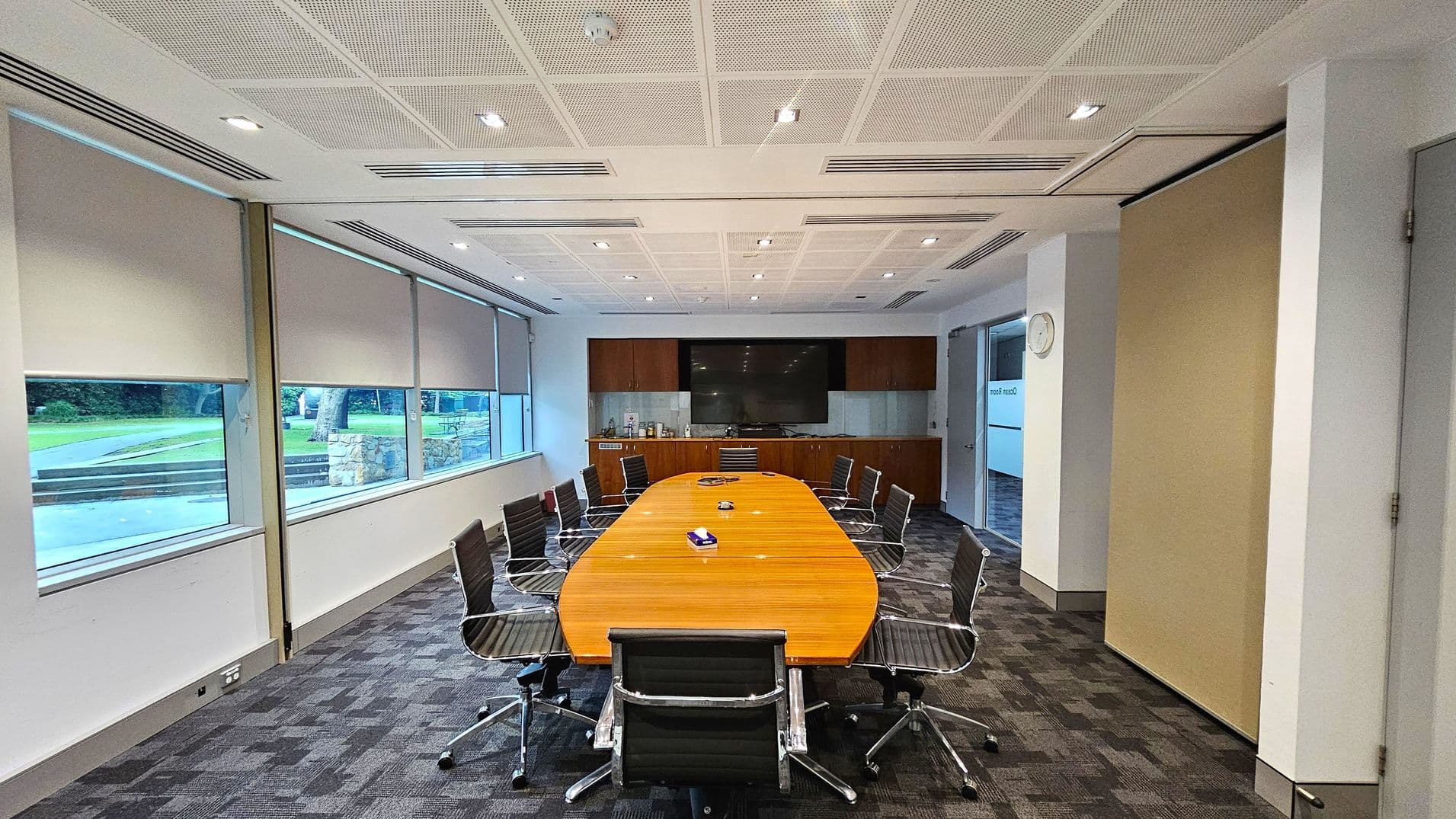 A conference room with a long, oval wooden table surrounded by black office chairs on a patterned carpet floor. The left side features large windows with roller blinds partially down, revealing a view of a grassy outdoor area. The back wall has a large flat-screen TV mounted above a wooden cabinet. The ceiling is white with recessed lighting. On the right, a wall clock is visible above a doorway.