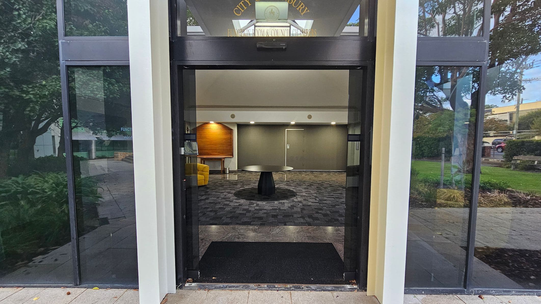 A glass entrance with a black mat on the floor leads into a building. Inside, there's a round black table on a patterned carpet. To the left, a yellow chair and a wooden board are visible. The walls are grey with a door in the centre. Outside, reflections of trees and greenery are seen on the glass panels. The ceiling inside is plain with recessed lighting.