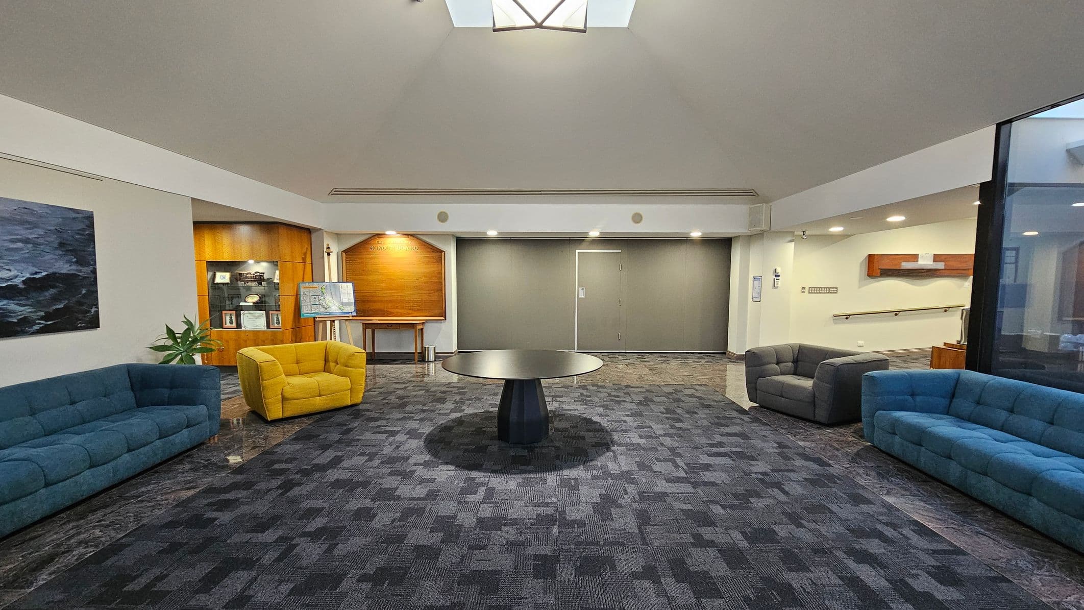 A spacious room with a patterned dark carpet. In the centre, a round black table stands on the floor. On the left, a blue sofa and a yellow armchair are placed near a wooden display case and a painting. On the right, another blue sofa and a grey armchair are positioned near a wall with a wooden shelf. The back wall features a wooden panel and a closed door. The ceiling is light-coloured with a skylight, and the room is well-lit with recessed lighting.