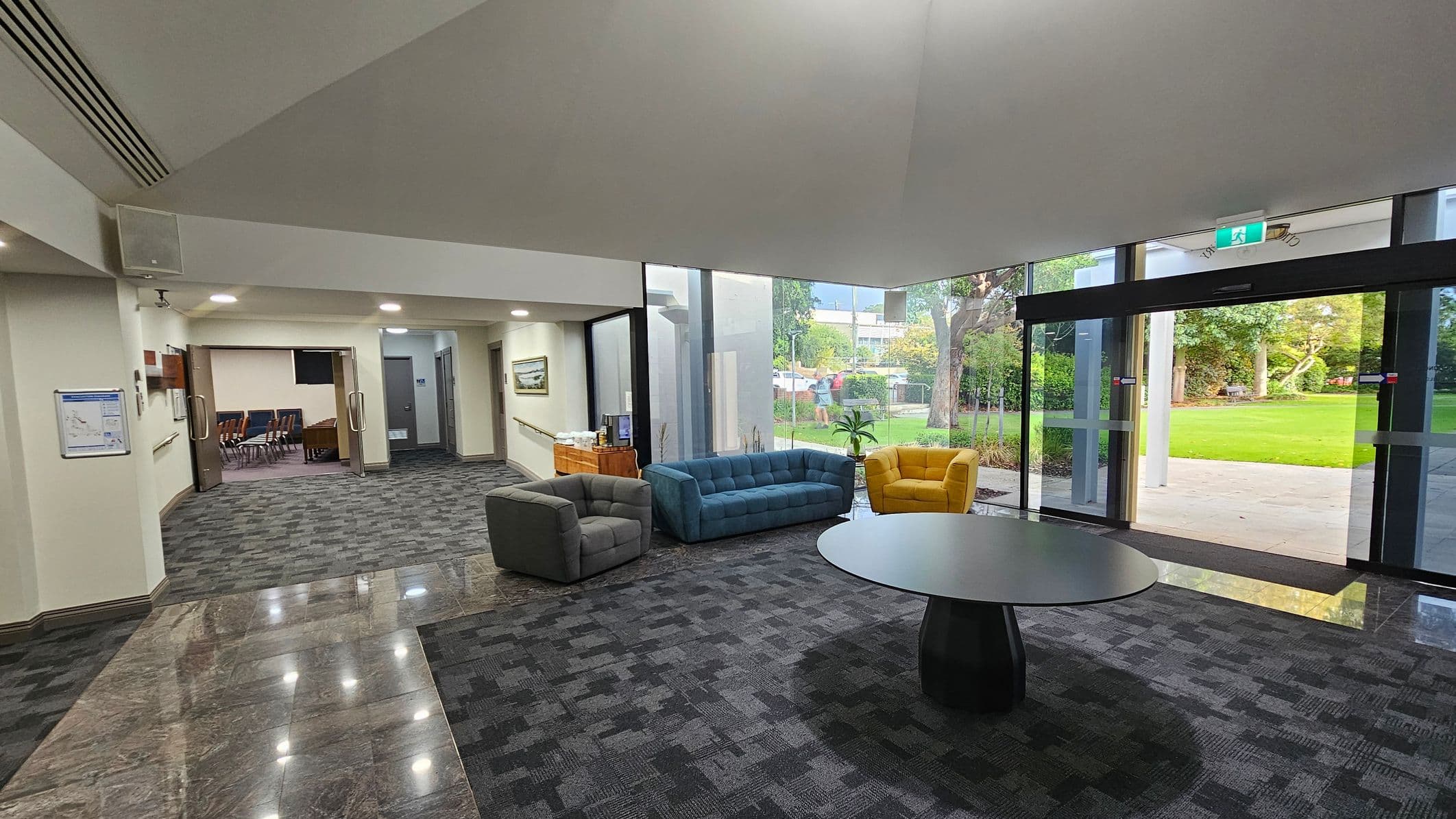 A spacious lobby area with a grey carpeted floor. On the left, there is an open doorway leading to a room with chairs. A grey armchair, a blue sofa, and a yellow armchair are arranged around a circular black table in the centre. Large glass doors on the right open to a green outdoor area with trees. The ceiling is high and light-coloured, with recessed lighting. A wooden cabinet with a coffee machine is against the wall near the glass doors.