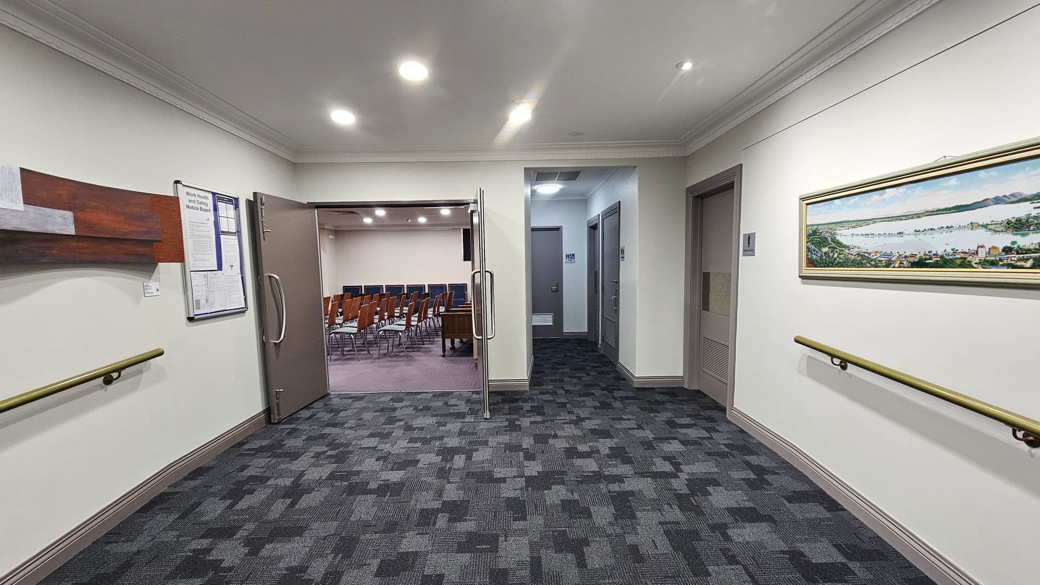 A hallway with a patterned carpet floor and white walls. On the left, there's a wooden notice board and a handrail. A door is open, leading to a room with rows of wooden chairs. On the right, a framed panoramic picture hangs above another handrail. Two closed doors are visible further down the hallway. The ceiling has recessed lighting.