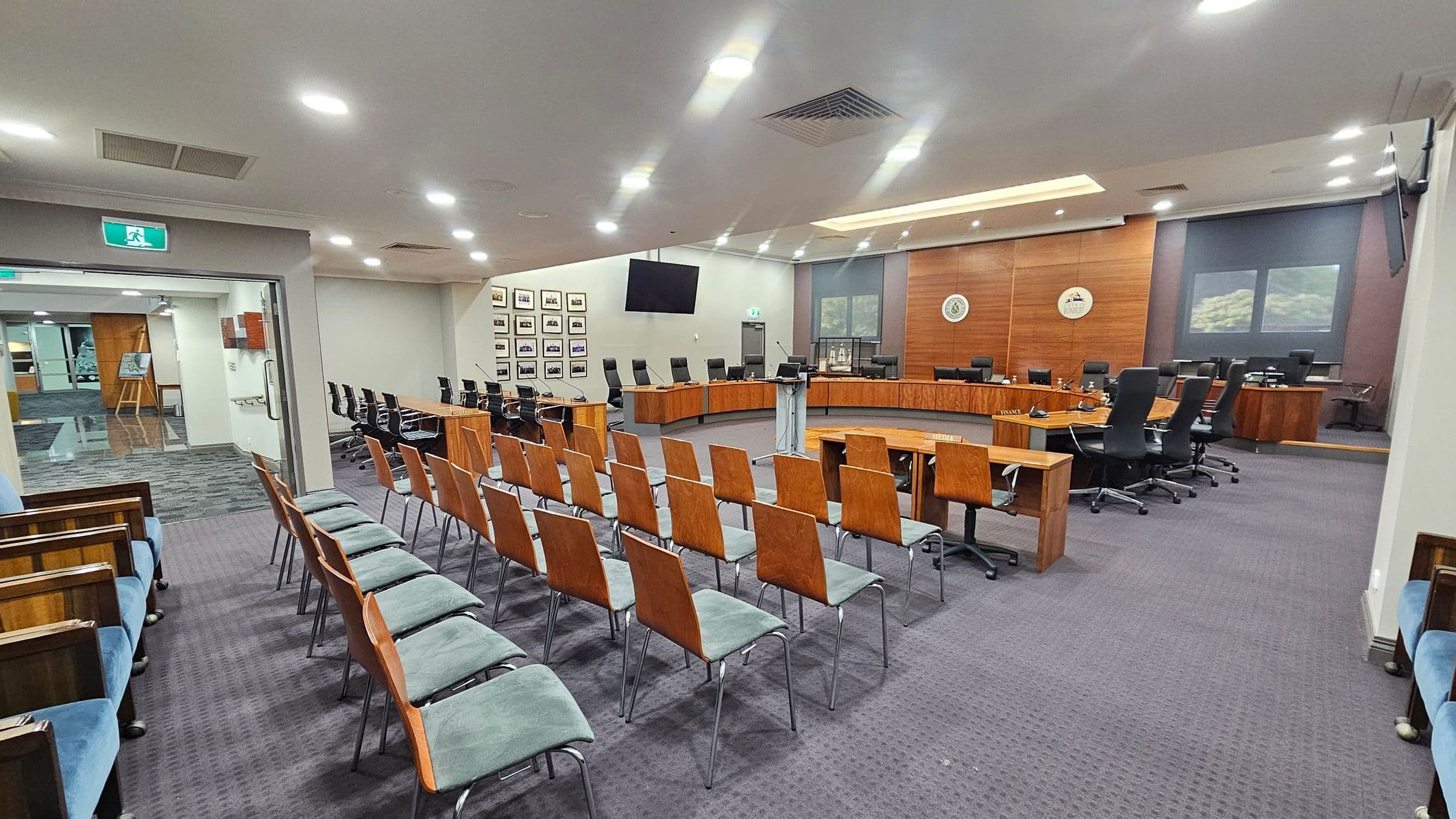 A spacious conference room with rows of wooden chairs with green cushions arranged in the foreground. To the right, a large U-shaped wooden table is surrounded by black swivel chairs. The back wall features wood panelling with two emblems and a large screen. On the left, there are framed pictures on the wall and an open doorway leading to another area. The ceiling is fitted with recessed lighting, and the floor is carpeted in a dark grey pattern.