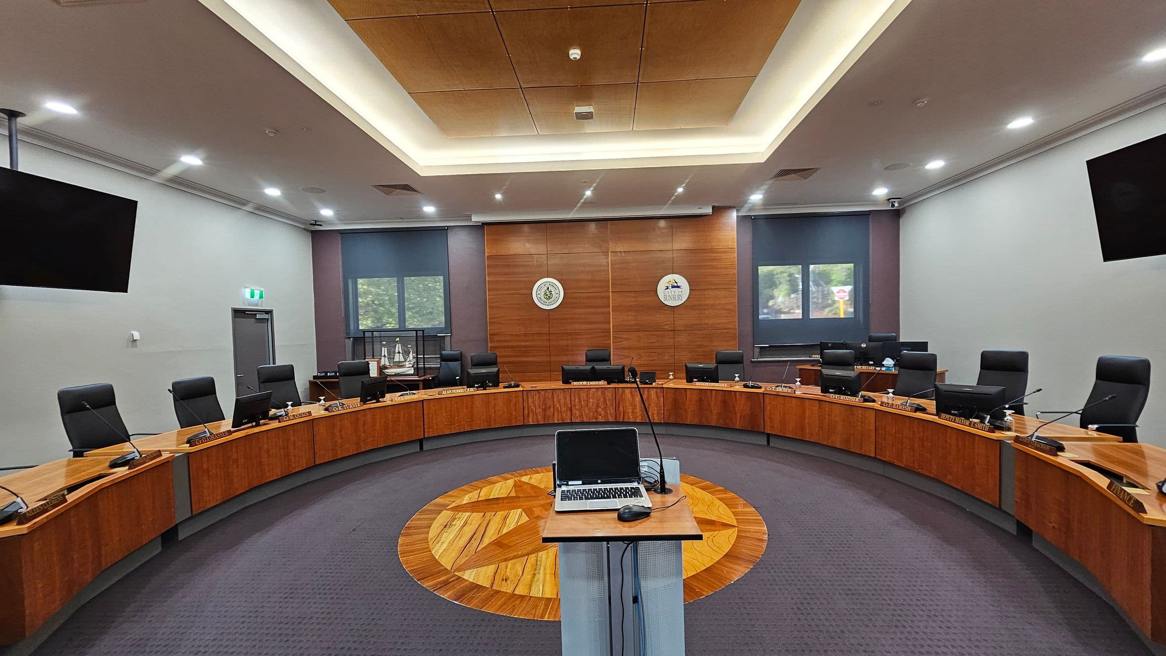 A modern conference room with a semi-circular arrangement of wooden desks and black chairs. In the centre, a podium with a laptop and microphone stands on a patterned wooden floor. The back wall features two emblems and two windows with blue blinds. The ceiling has recessed lighting and a central wooden panel. Two large screens are mounted on the side walls. The floor is carpeted in grey.