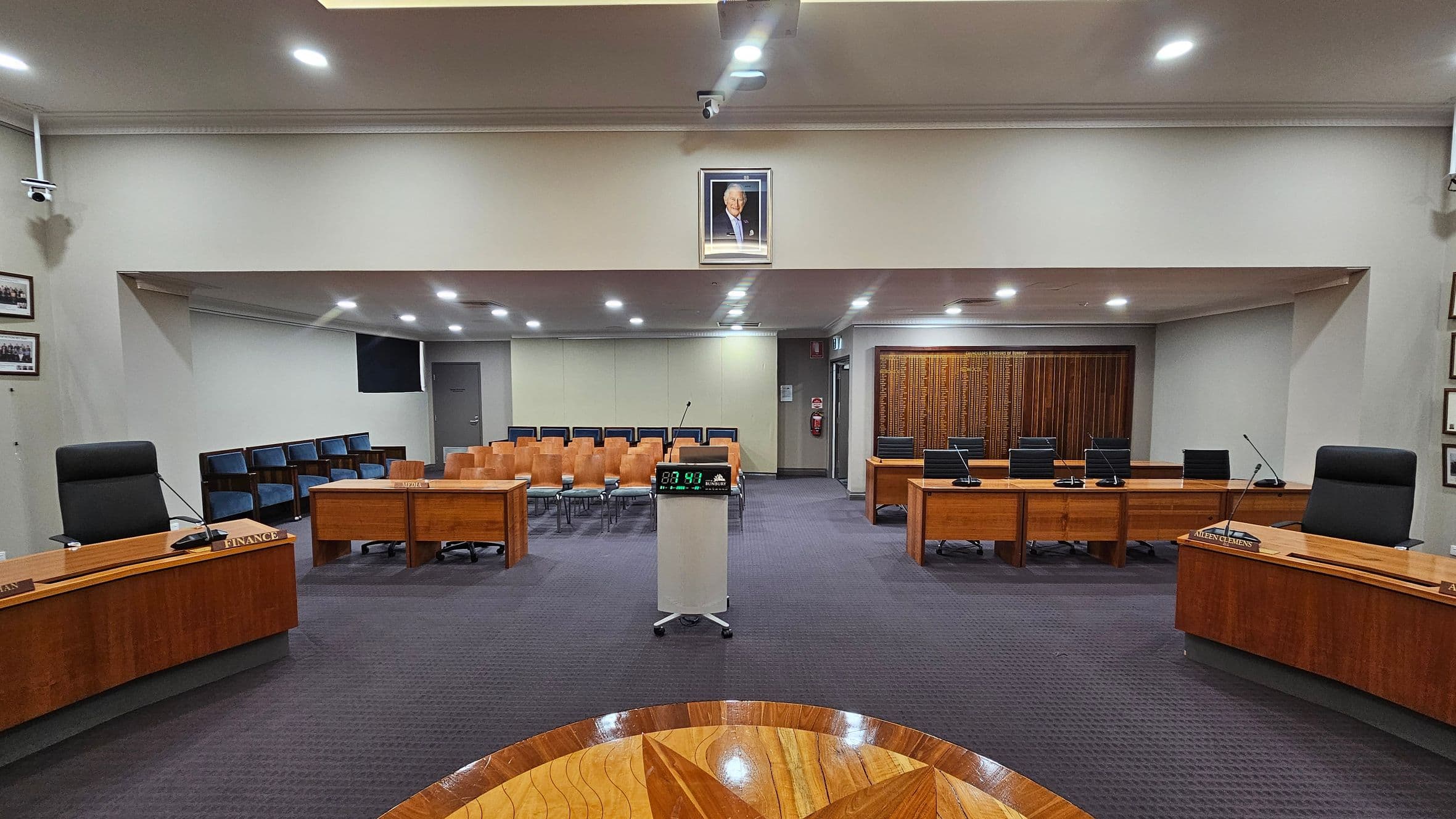 A council chamber with a central podium featuring a digital timer. Surrounding the podium are wooden desks with microphones and nameplates. On the left and right are black chairs behind the desks. In the background, rows of wooden and blue chairs face the front. A framed portrait hangs above the central area. The floor is carpeted in dark grey, and the ceiling has recessed lighting. Walls are adorned with framed photos and a wooden panel.