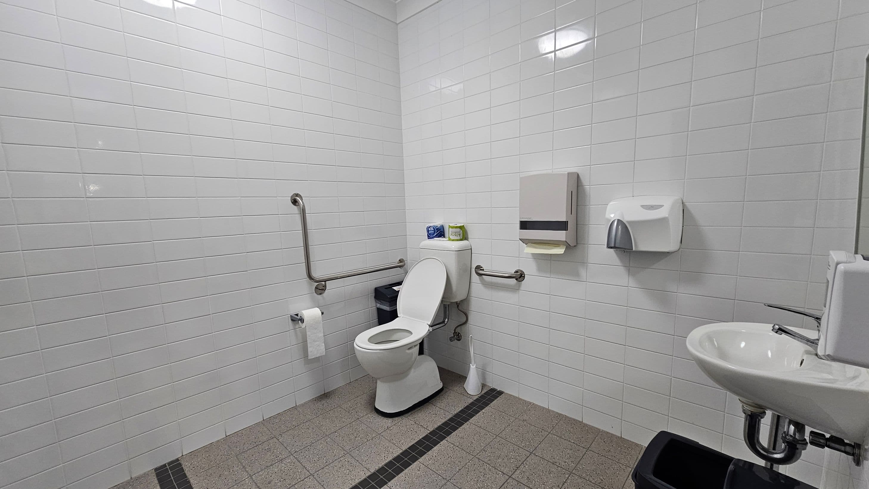 A public restroom with white tiled walls and a grey tiled floor. On the left, there is a toilet with a raised seat, equipped with grab bars on both sides. Above the toilet, there are two rolls of toilet paper. A toilet brush is on the floor beside it. On the right wall, there is a paper towel dispenser and a soap dispenser above a small sink. Below the sink, a black waste bin is visible. The ceiling has a simple light fixture.