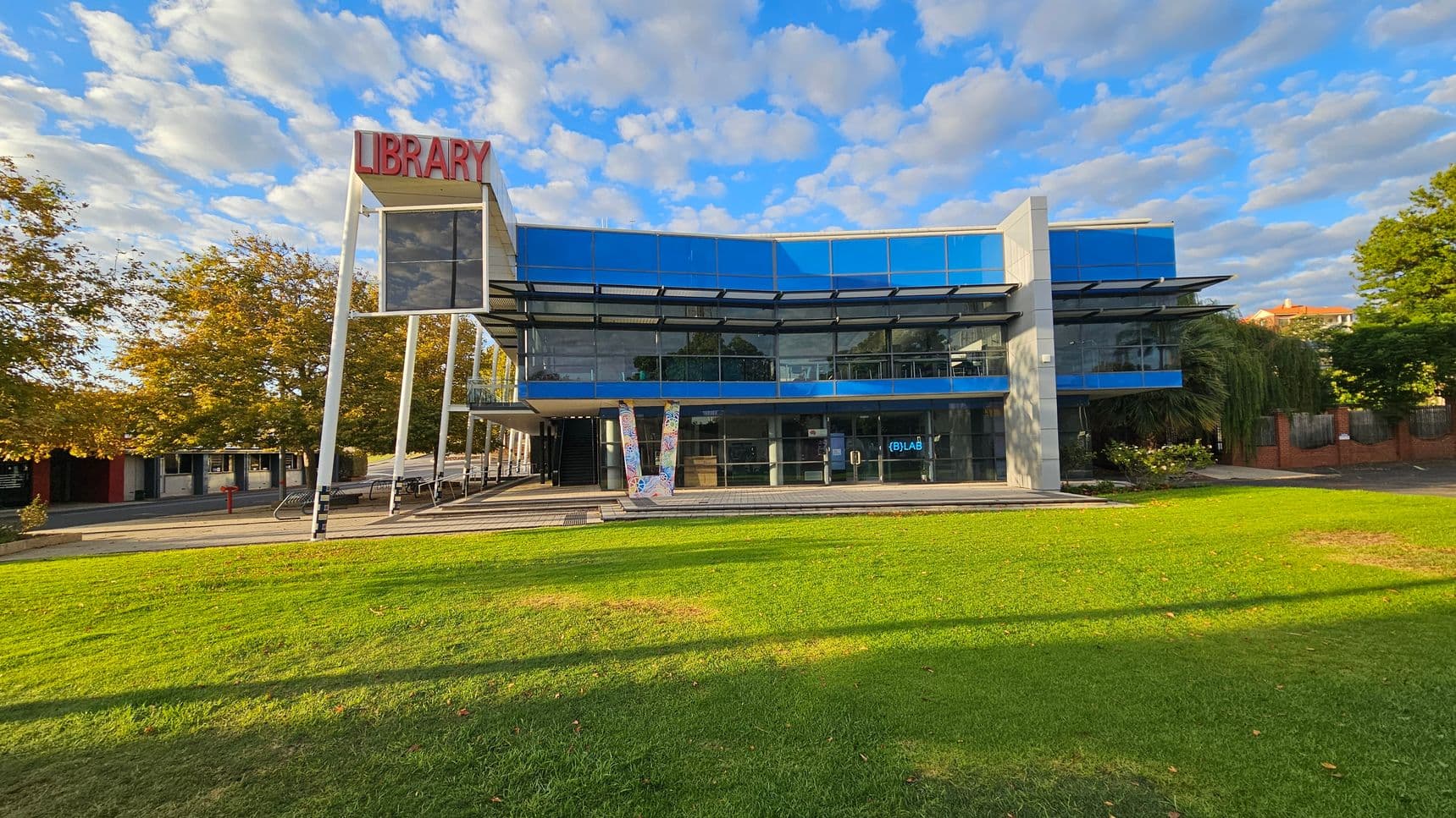 A modern library building with a glass facade and a prominent red "LIBRARY" sign on the left. The structure features large windows and a blue-tinted upper section. In front, there's a well-maintained green lawn. To the left, there are trees with autumn foliage and a few benches. The right side has more greenery and a brick wall. The sky is partly cloudy, casting shadows on the grass.