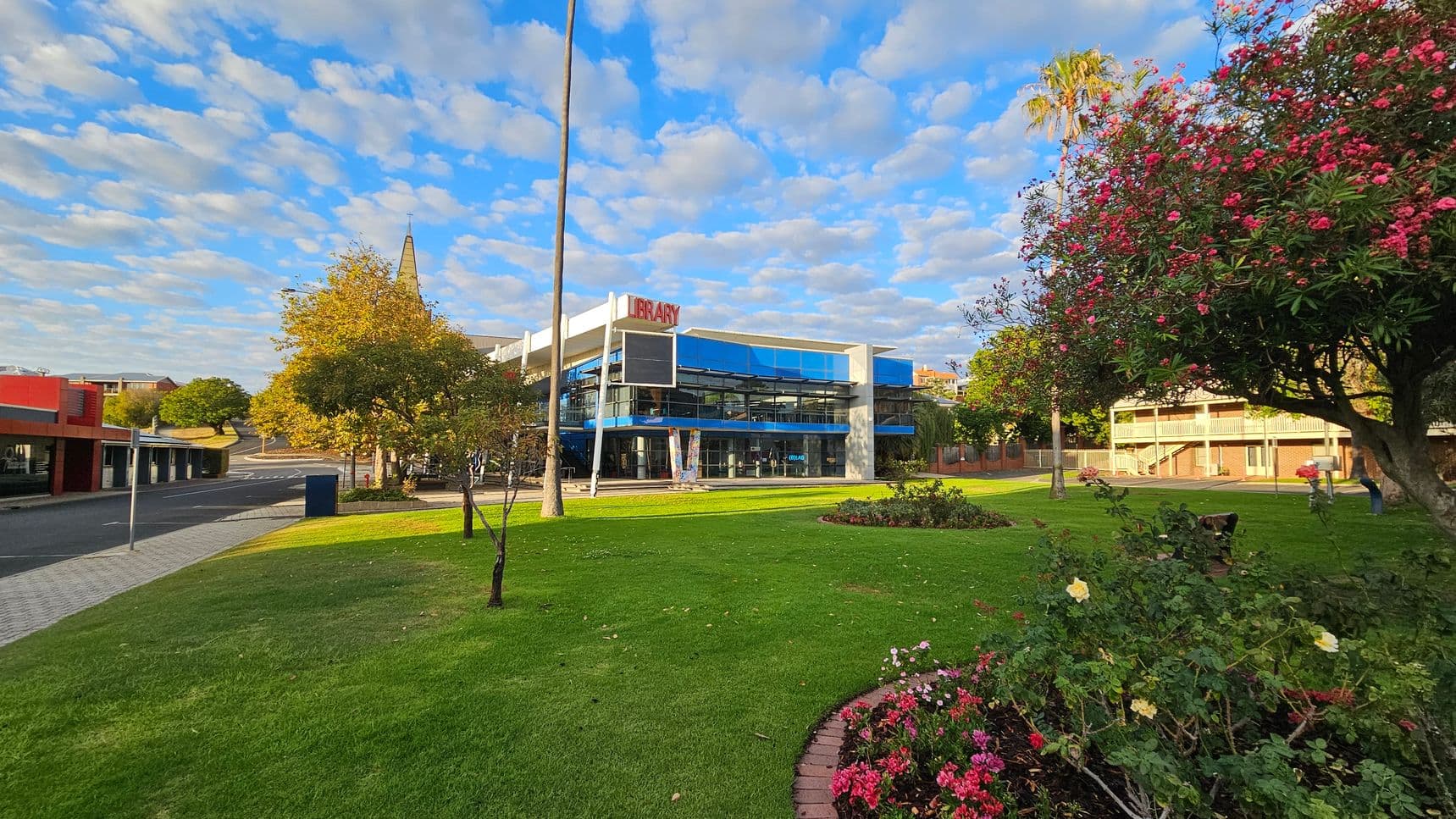A modern library building with a glass facade is in the centre, surrounded by a well-maintained green lawn. On the right, there are trees with pink flowers and a flower bed with various blooms. To the left, a paved walkway runs alongside a road with a red and grey building. In the background, a tall spire is visible, and a palm tree stands near the library. The scene is well-lit, suggesting daytime.
